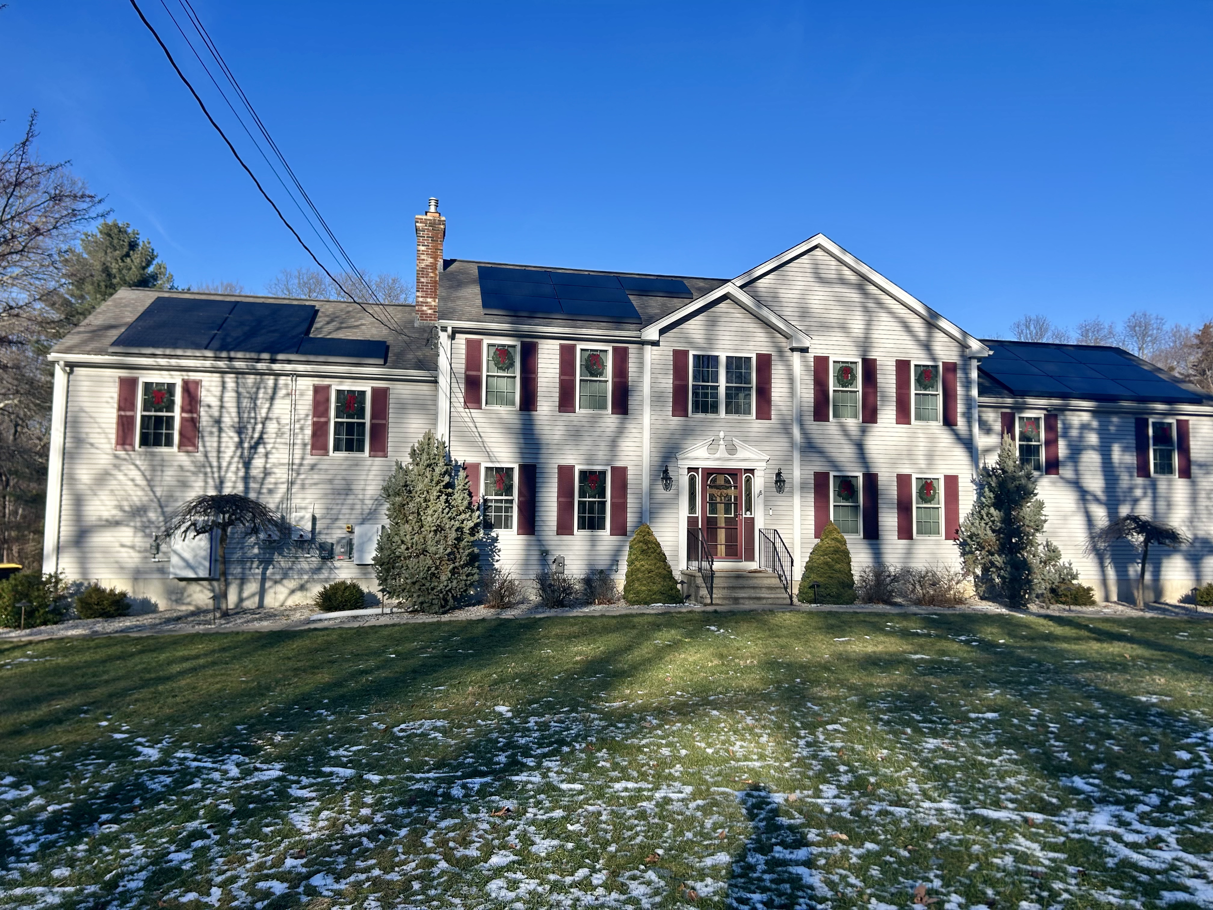A large two-story house with white siding, red shutters, and a brick chimney. Solar panels are on the roof, and holiday wreaths are on the windows. The front yard has green grass with some patches of snow, small bushes, and trees with shadows cast across the house. The sky is clear and blue.