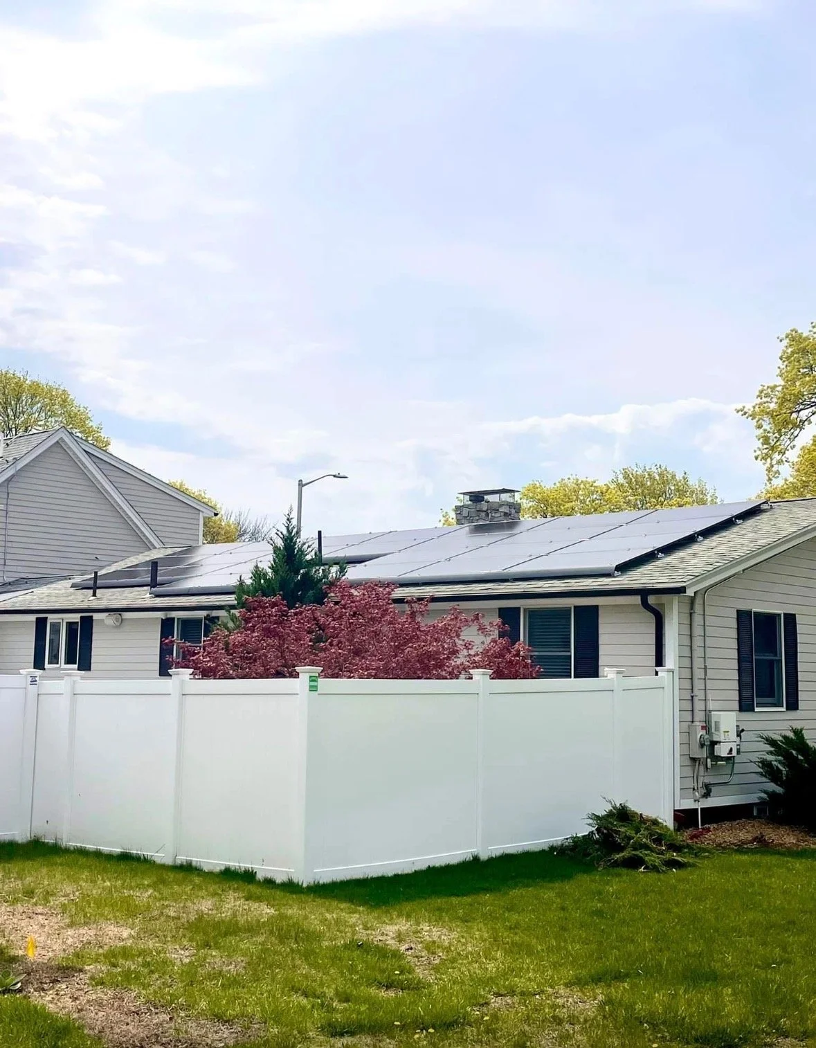 A house with solar panels on the roof, a white vinyl fence, a pink flowering tree, and green grass, under a partly cloudy sky.