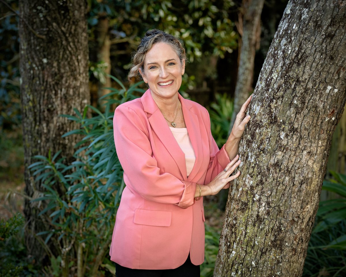 Teresa Roberts, leadership coach and co-founder of Secure On Every Front, smiling outdoors in a coral blazer leaning against a tree.