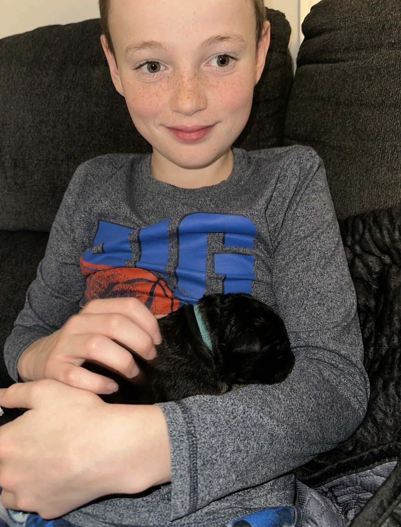A boy with red hair and freckles sitting on a couch, holding a small black puppy with a blue collar.