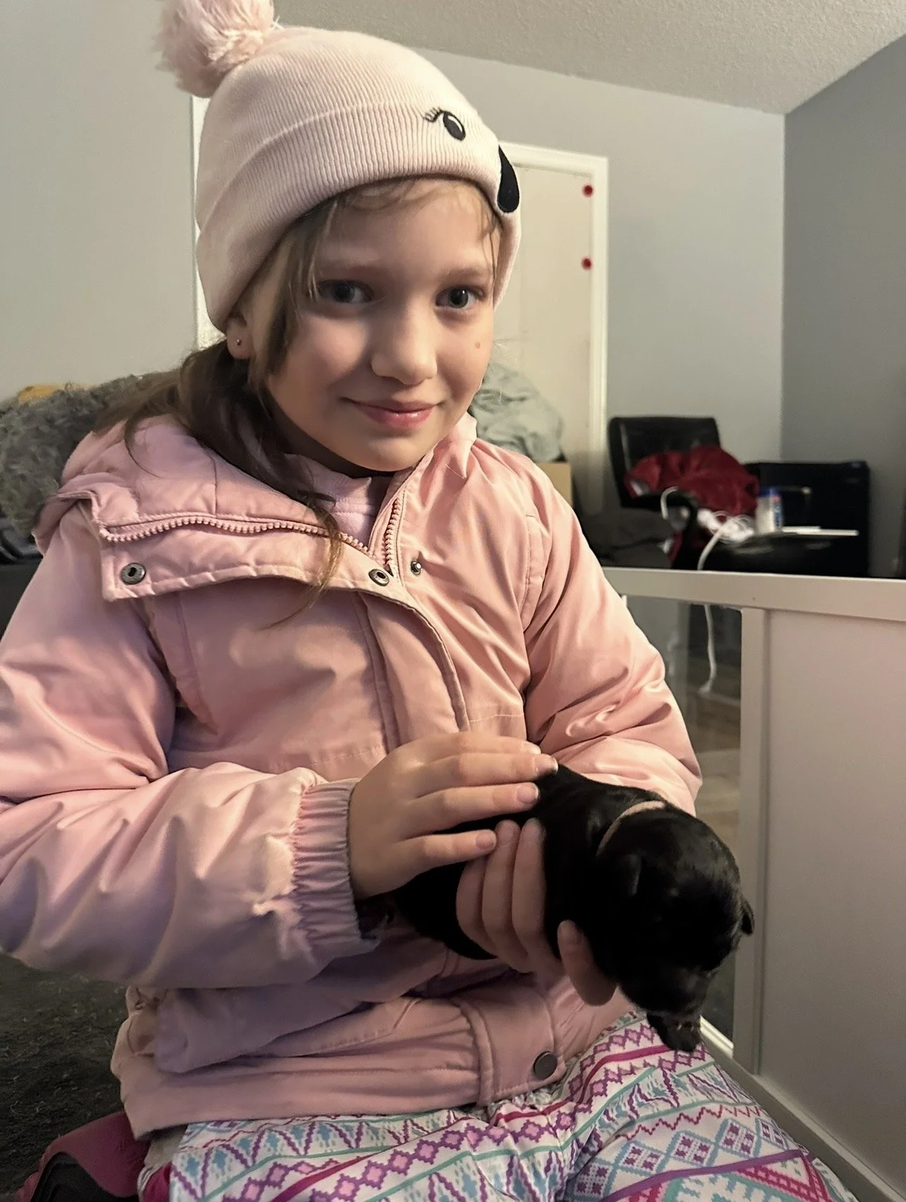 A young girl wearing a pink winter jacket and a pink knit hat with a dog face design, holding a small black puppy indoors.