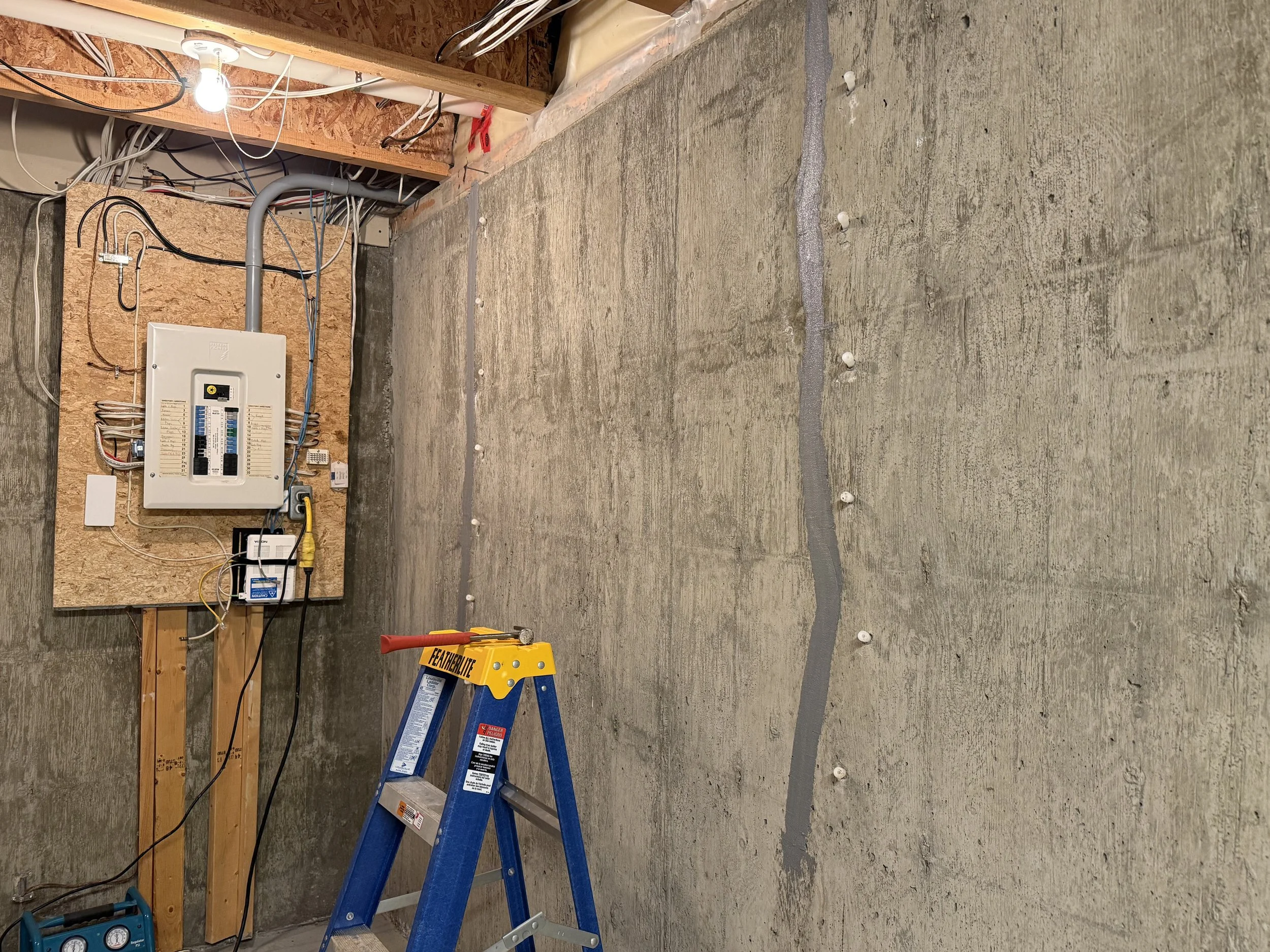 Interior of a basement with concrete walls and floor, a step ladder, power tools, a tool bag, and electrical panels.  Too long cracks on the foundation have just been sealed.
