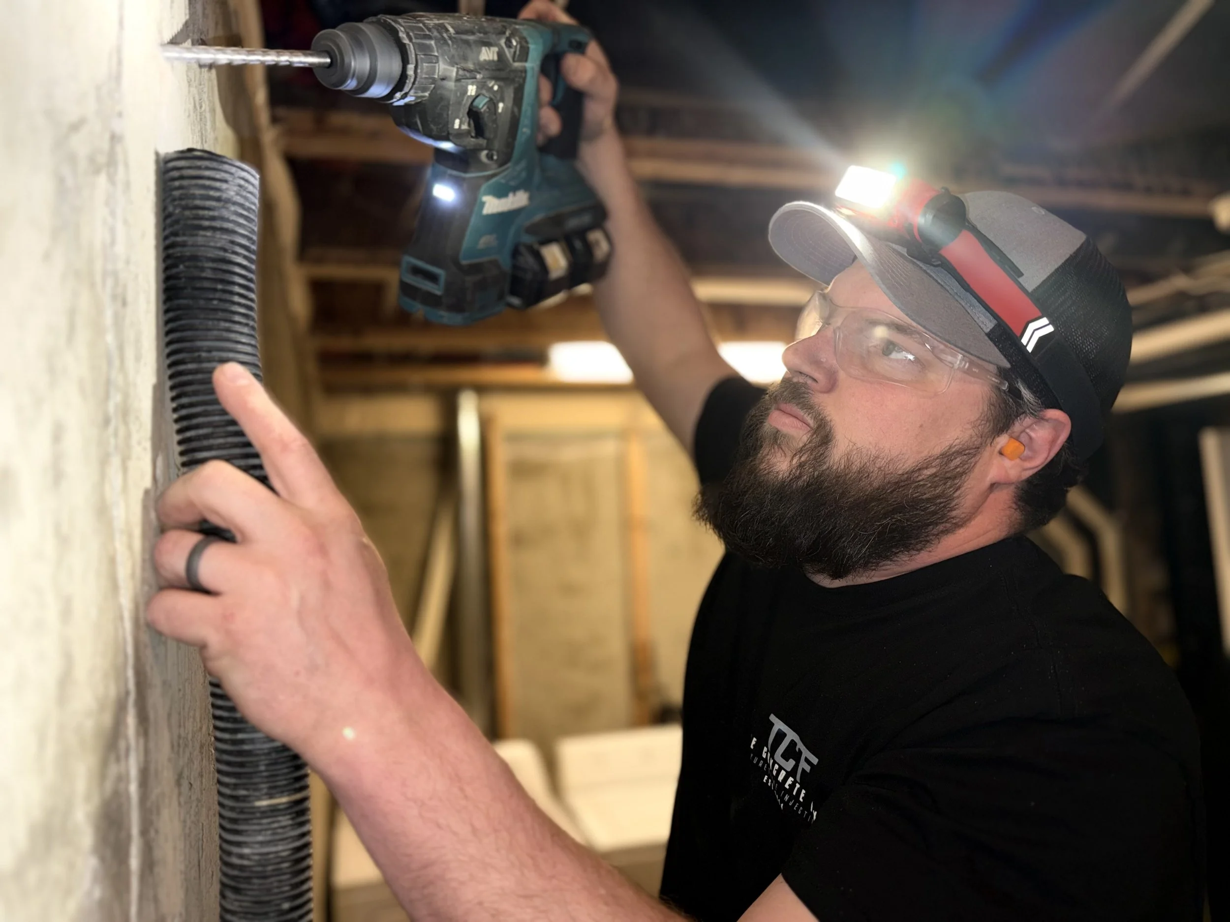 Man wearing safety glasses, a cap with a headlamp, and hearing protection drilling a hole in a foundation using black tube vacuum to collect the dust.