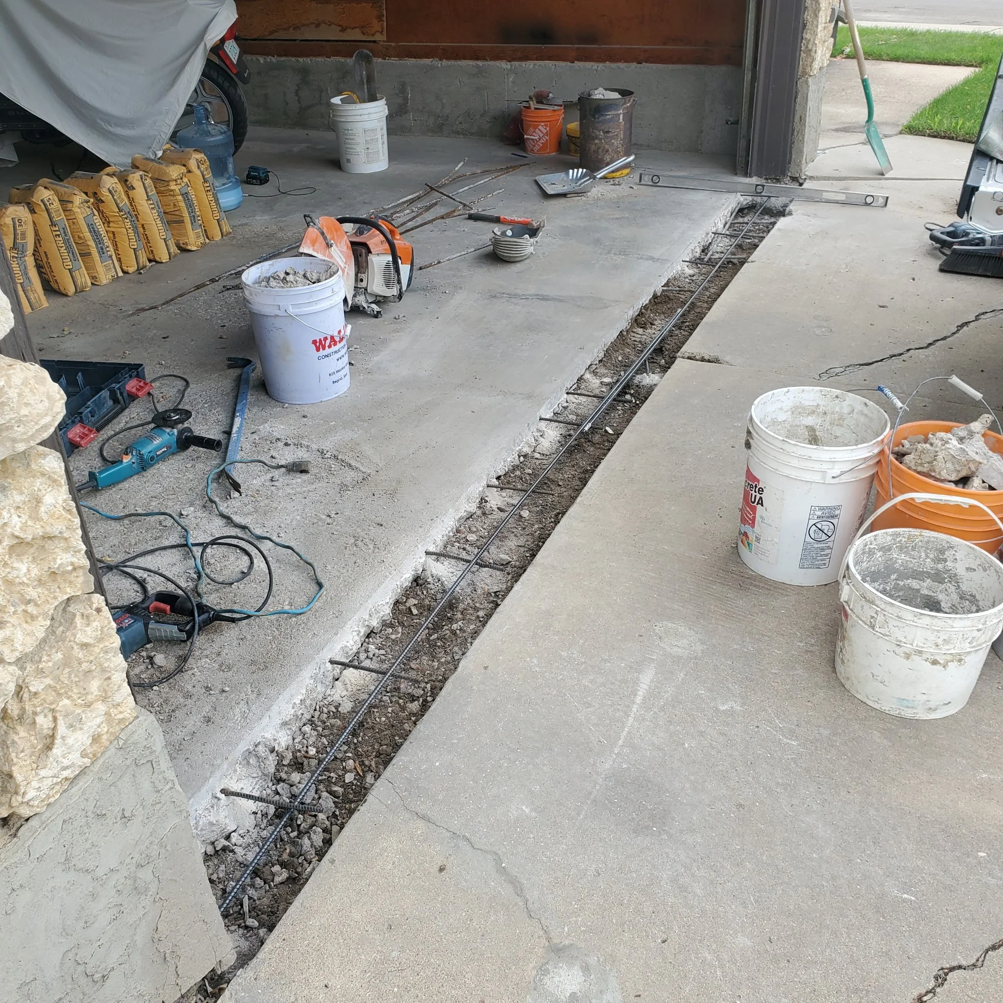 Construction work on a concrete driveway, with tools and buckets of materials, and rebar laid in the trench.