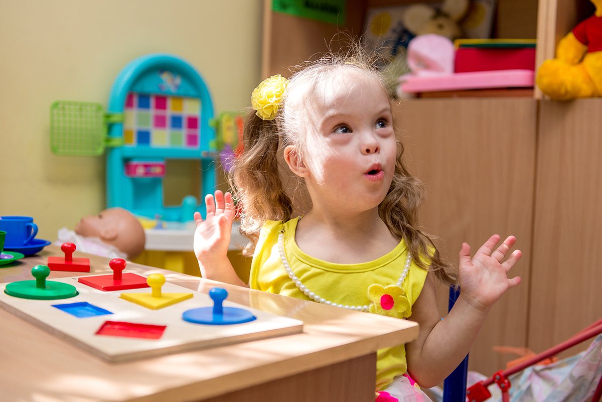 A young girl wearing a yellow shirt and a yellow flower hair clip, sitting at a table with colorful game pieces, with a playful expression and hands raised working on language skills during speech therapy for children in Norwalk, CT.