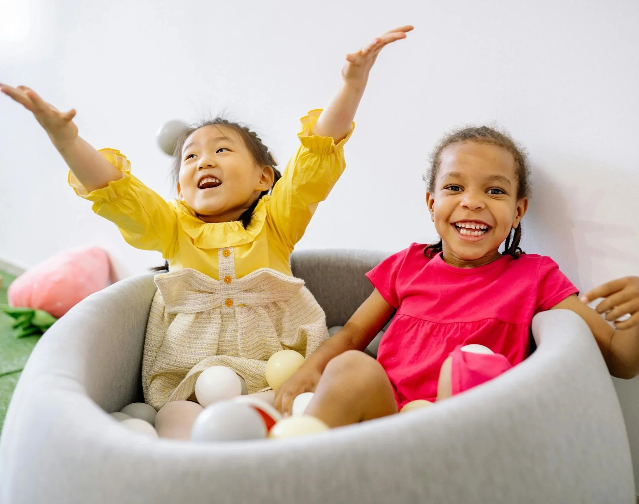 Two young girls playing in a ball pit, smiling and laughing. One girl is wearing a yellow dress with ruffled sleeves, the other in a pink shirt. They are working on communication during play-based speech therapy in Norwalk, CT.