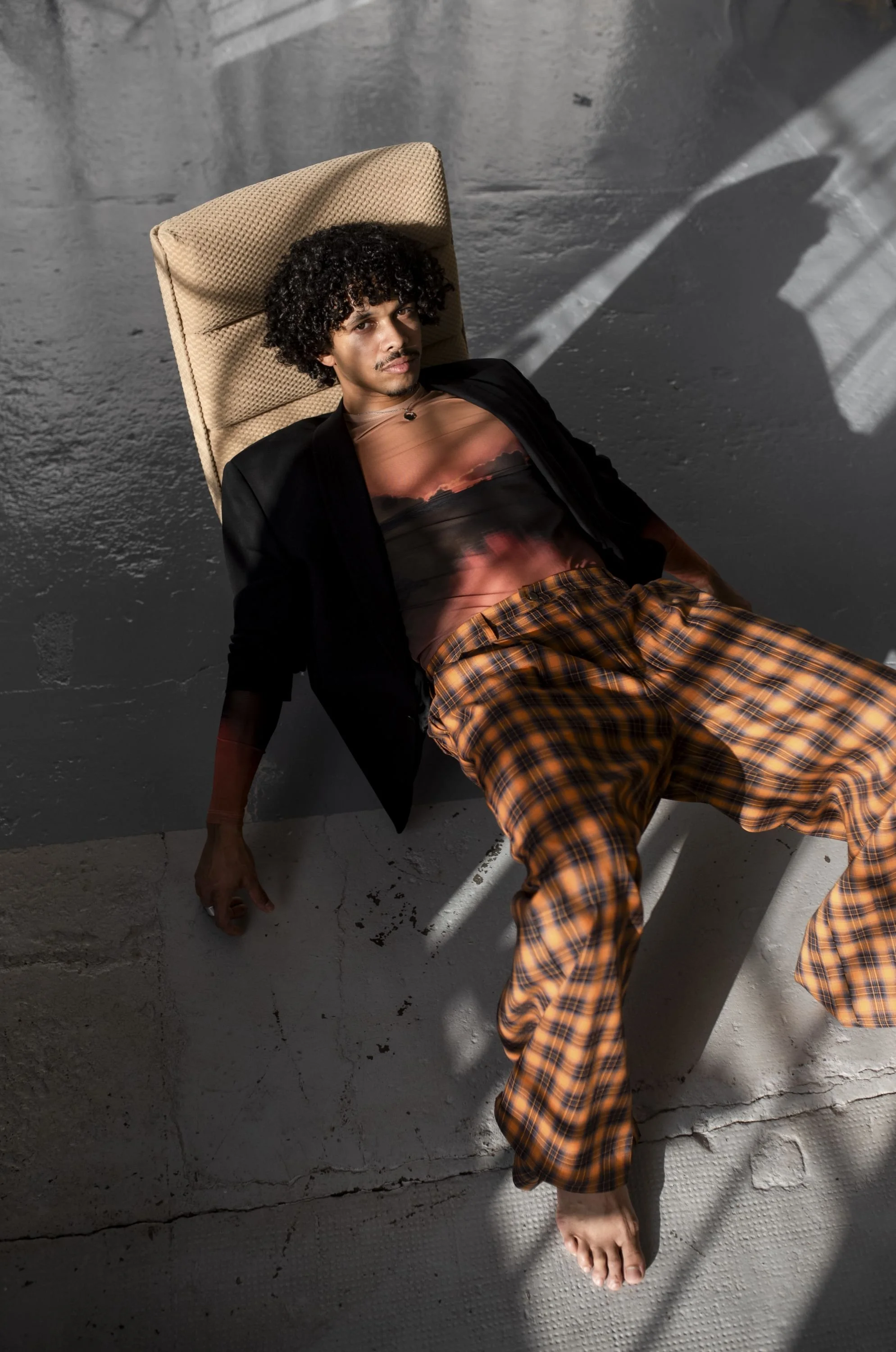A young man with curly hair is lying on the floor, wearing a black blazer and orange plaid pajama pants, with a sunset sky graphic shirt. He is leaning against a beige chair in a room with gray concrete walls and floor, with sunlight casting shadows across the scene.