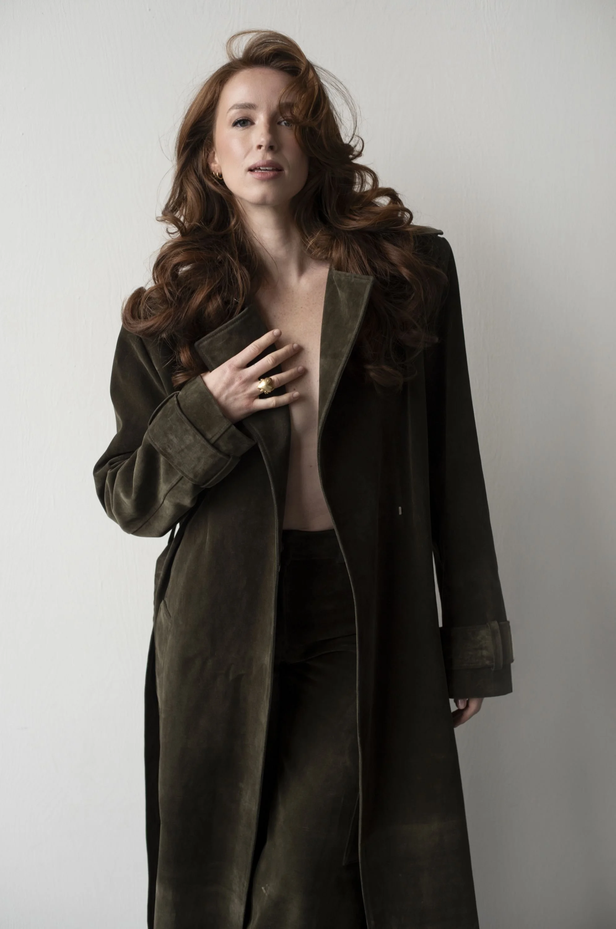A woman with long, curly red hair wearing a dark green coat, partially open, revealing her bare chest, standing against a plain light background.