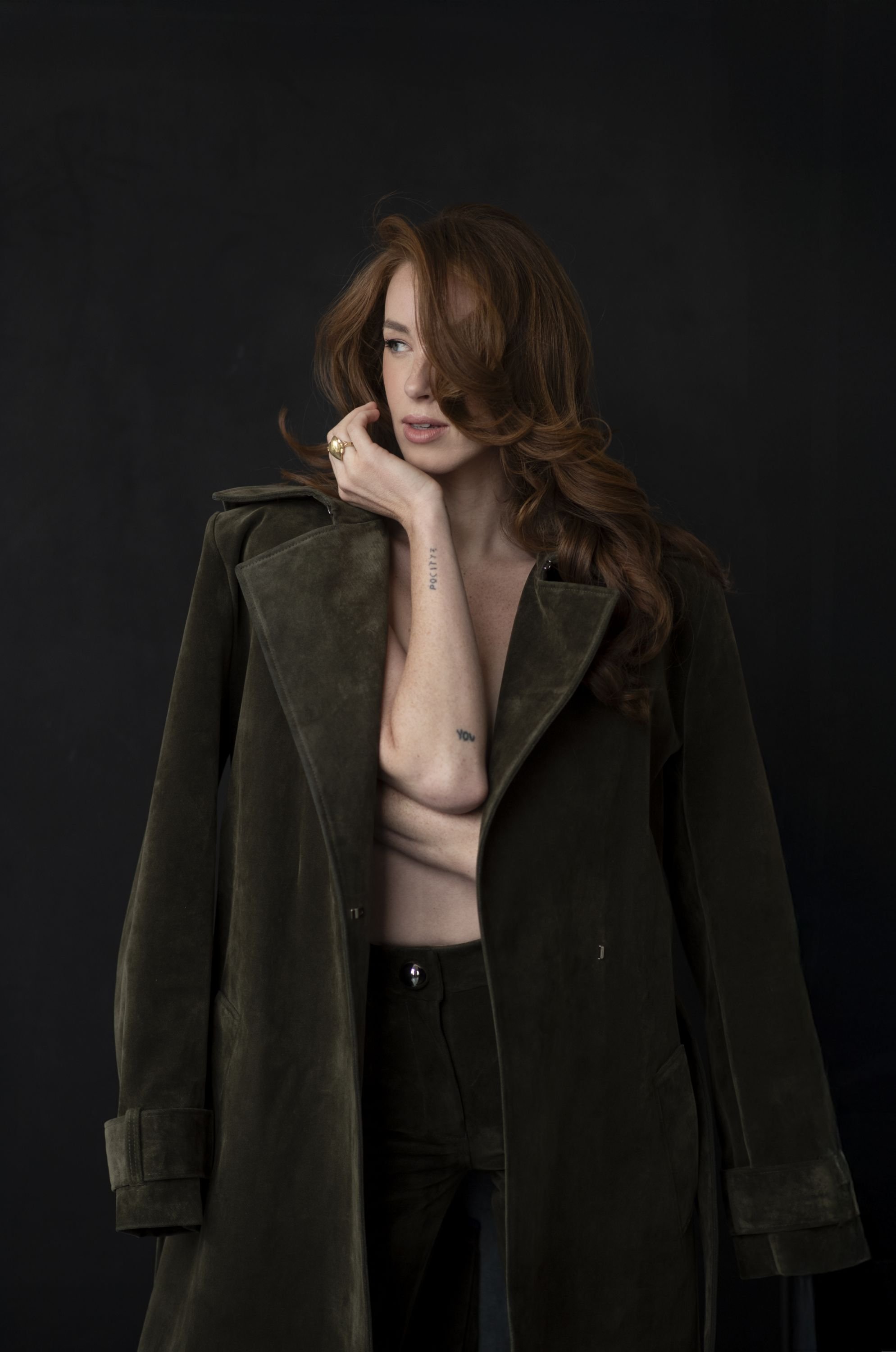 A woman with red hair, partially topless, wearing a dark green trench coat over black pants, standing against a plain black background.