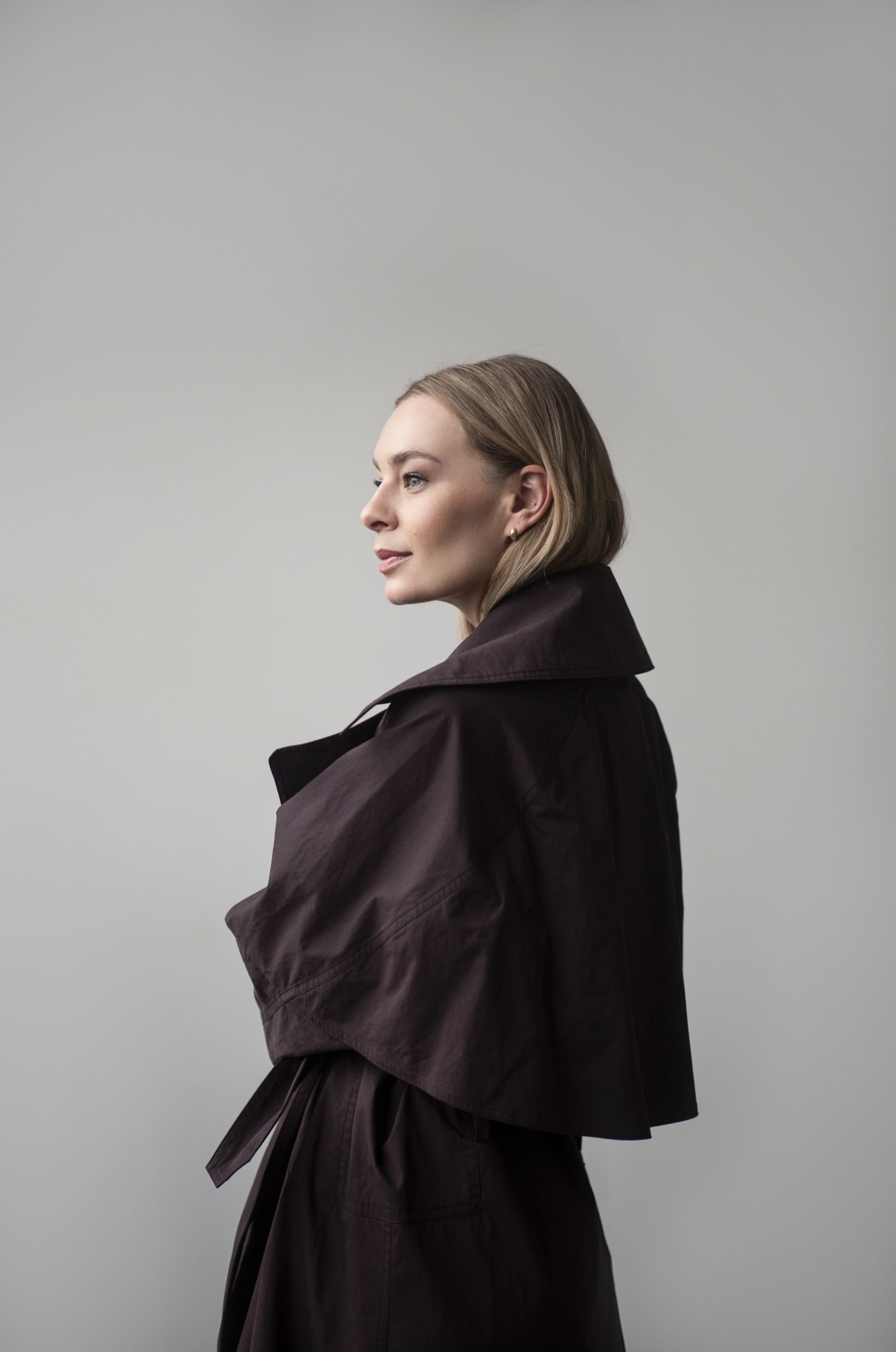 Side view of a woman with short blonde hair wearing a dark trench coat against a plain grey background.