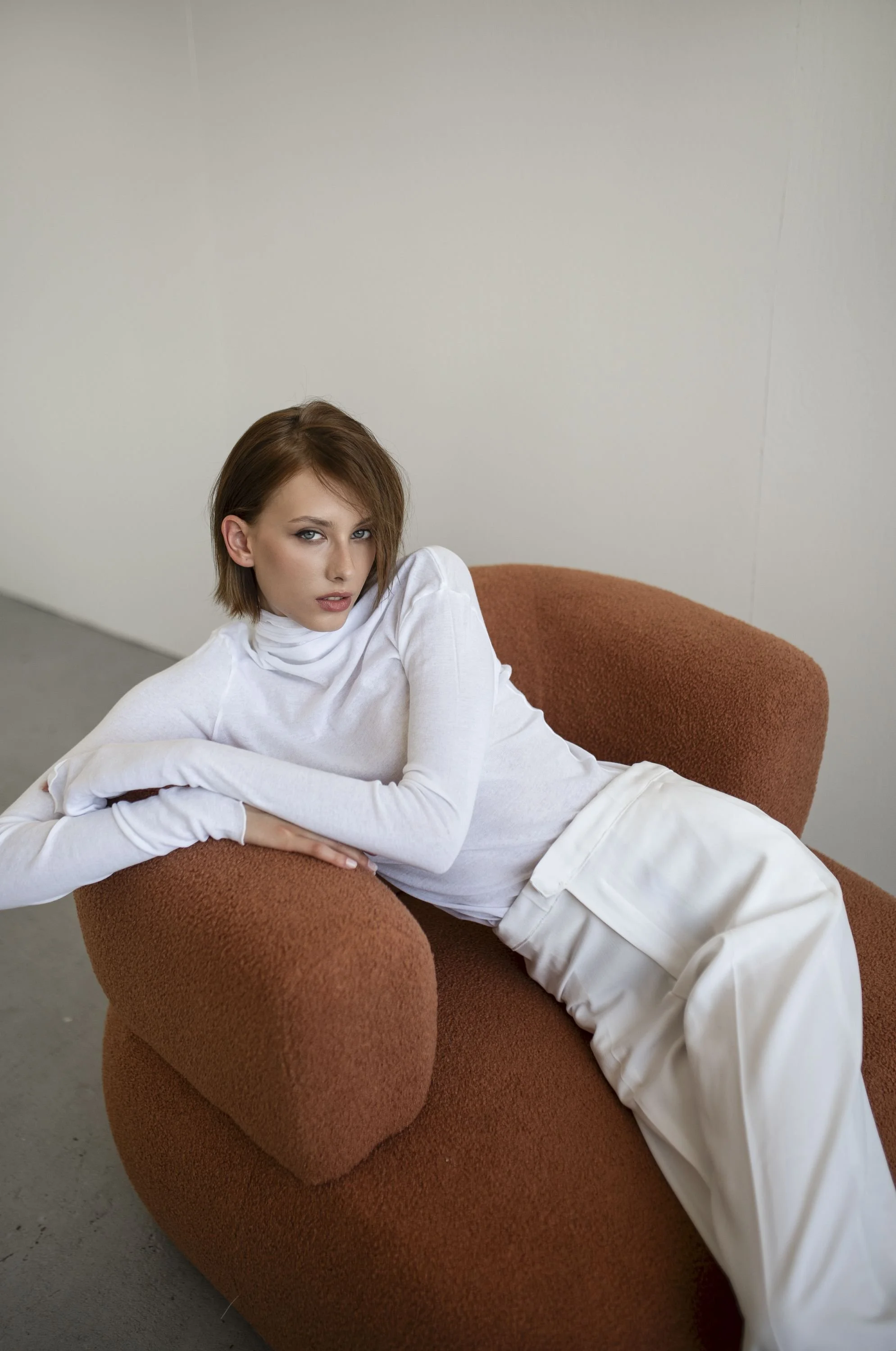 A woman with short brown hair and blue eyes lounging on a reddish-brown textured sofa against a plain white wall, wearing a white long-sleeve turtleneck and cream-colored pants.