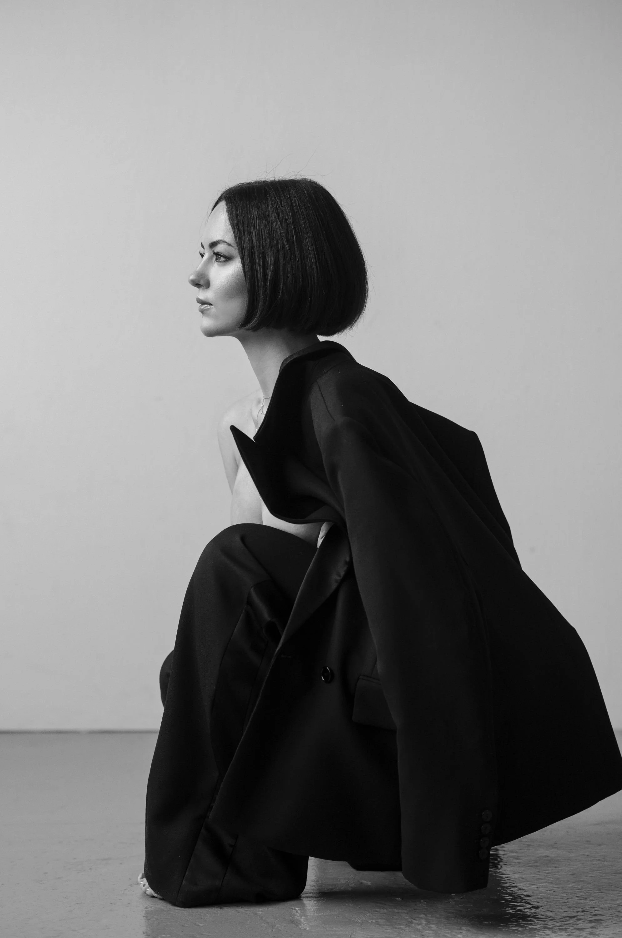 A black and white photo of a woman with a short bob haircut, sitting on the ground with her legs folded, wearing a large dark blazer draped over her shoulder, looking to the left.