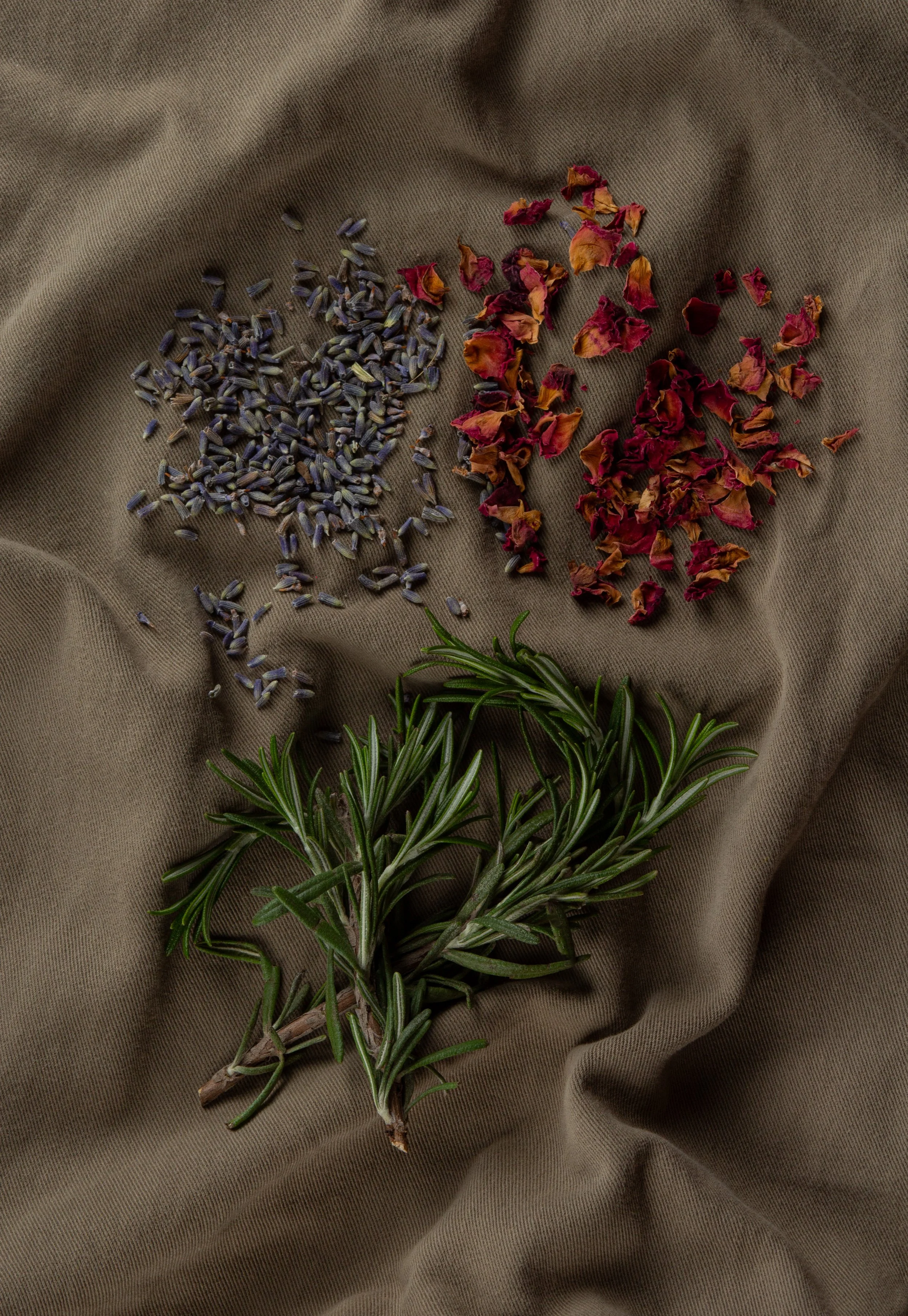 Dried lavender buds, dried rose petals, and fresh rosemary on a brown fabric background.