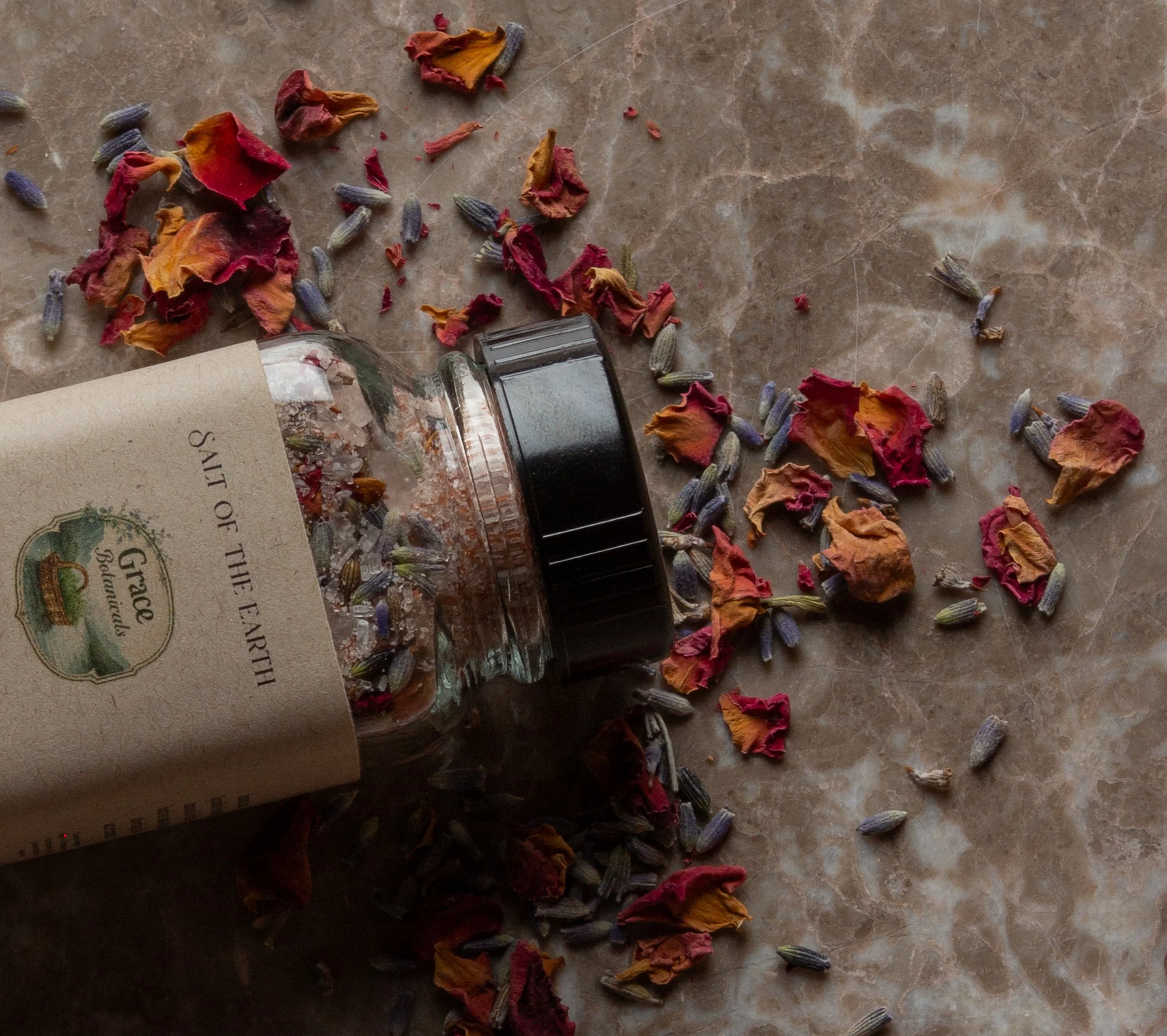 An overturned glass jar labeled 'SIT OF THE EARTH' with dried flower petals and lavender scattered on a marble surface.