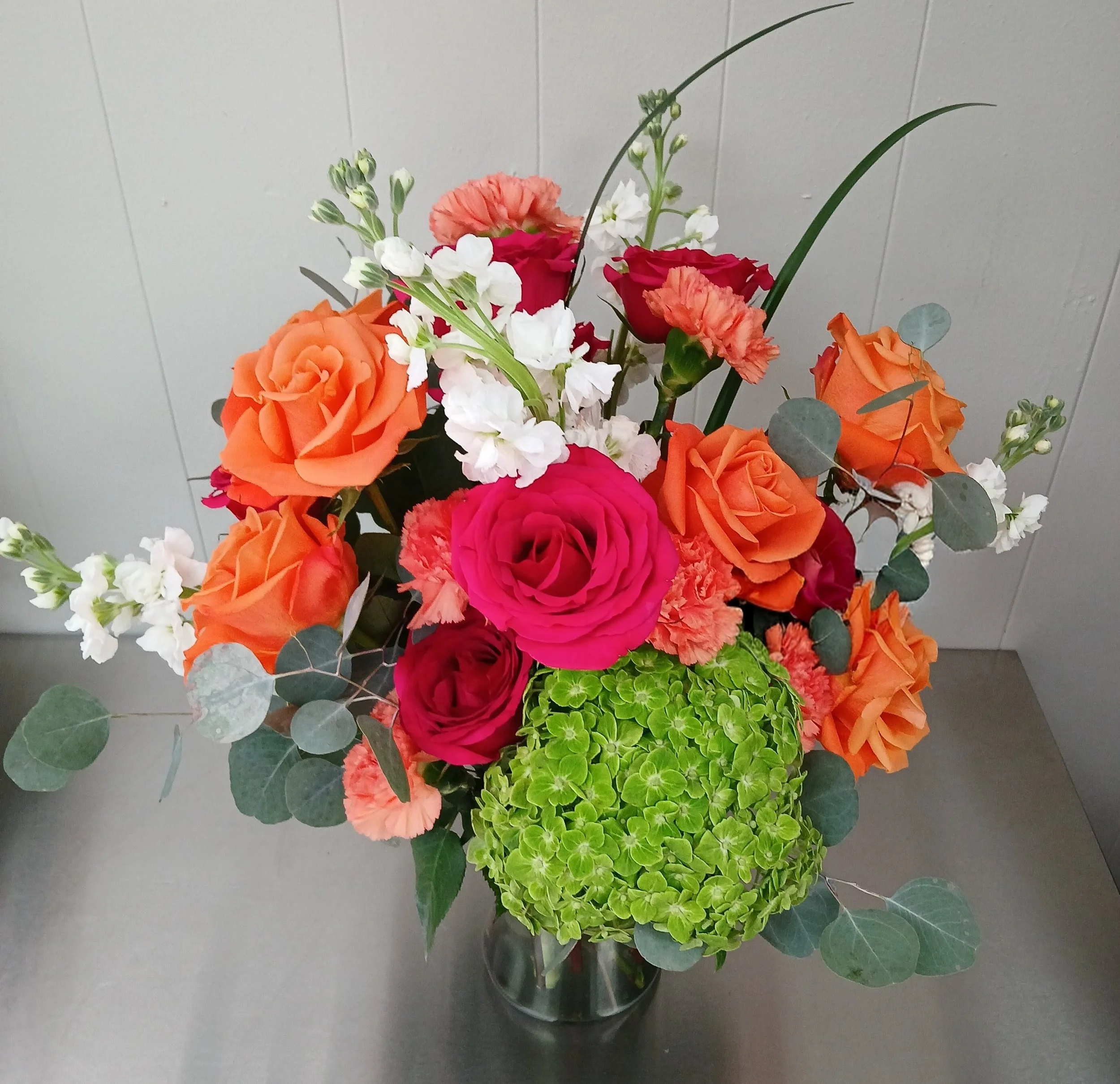 Colorful flower arrangement with orange, pink, and white roses, green hydrangea, white stock flowers, and eucalyptus leaves in a glass vase.