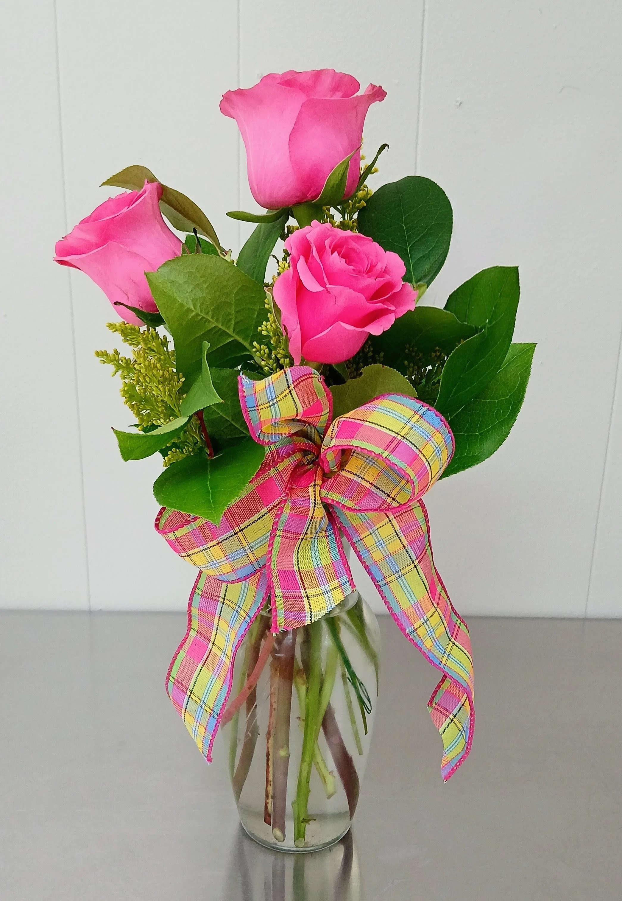 A small glass vase with three pink roses, green leaves, and a plaid ribbon tied in a bow, on a gray surface against a plain white background.