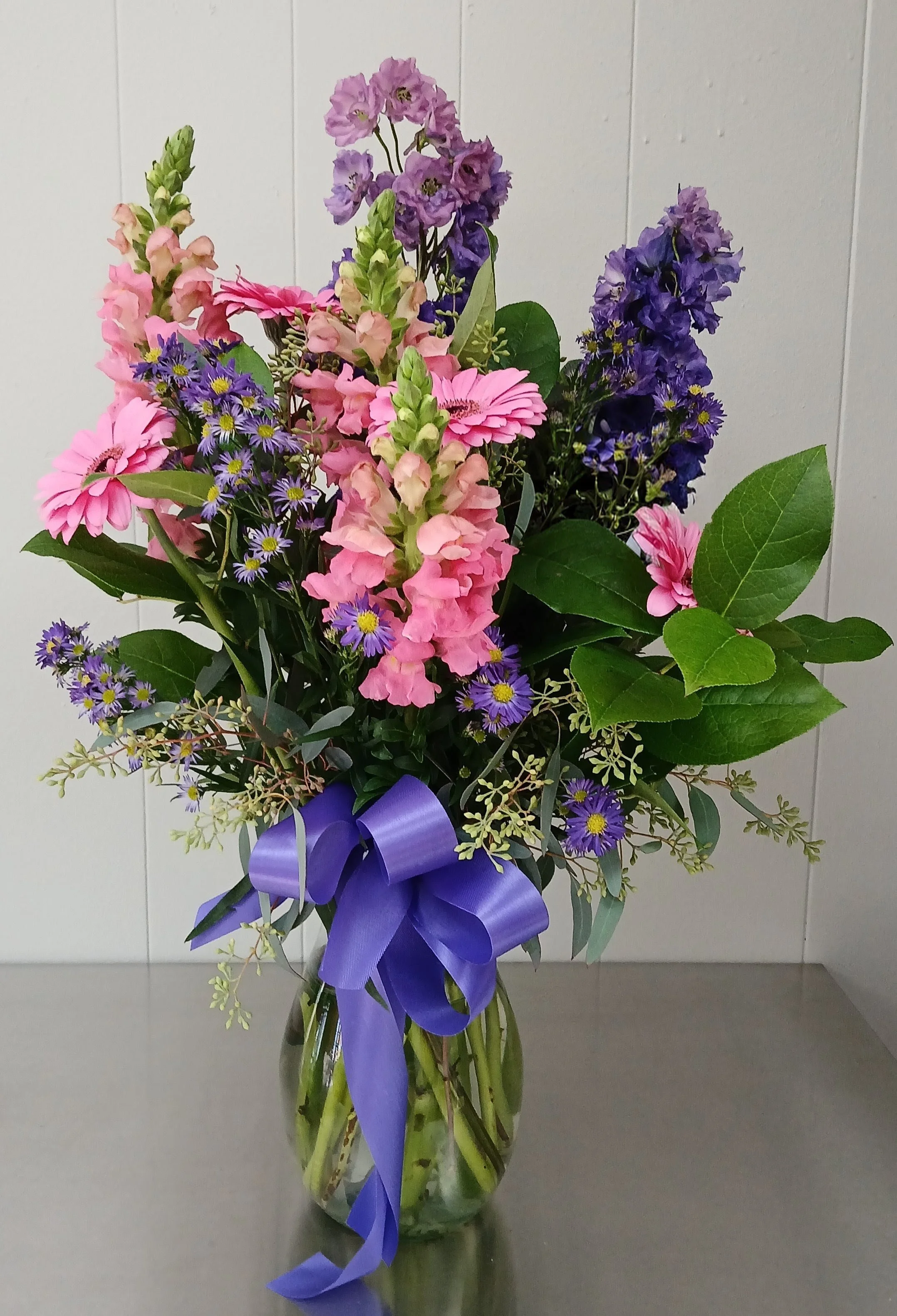 A bouquet of pink, purple, and blue flowers in a glass vase with a purple ribbon tied around it.