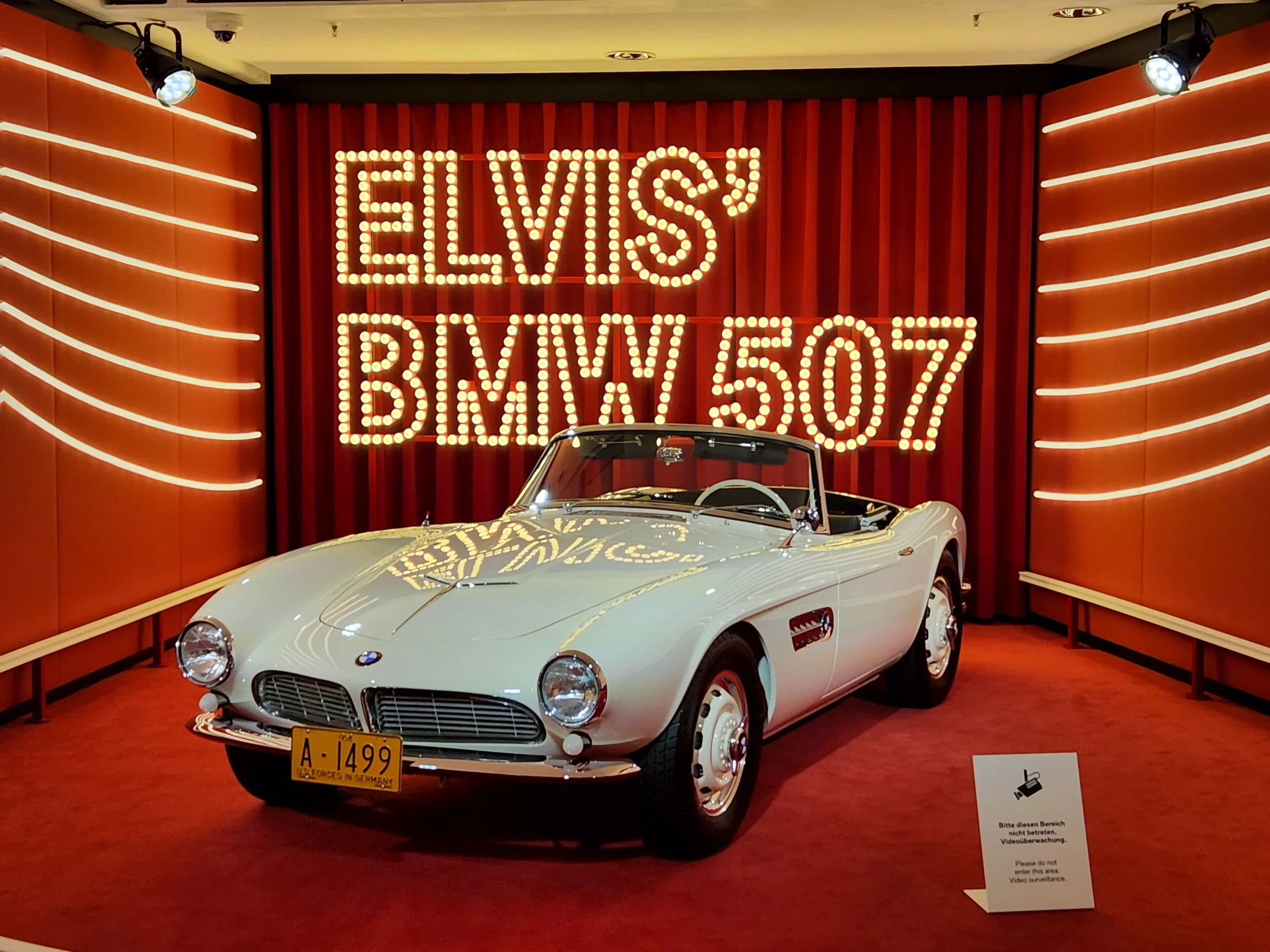 A vintage white BMW 507 convertible car displayed in a museum or showroom with red curtains and marquee sign in the background reading "Elvis BMW 507" in bright lights.