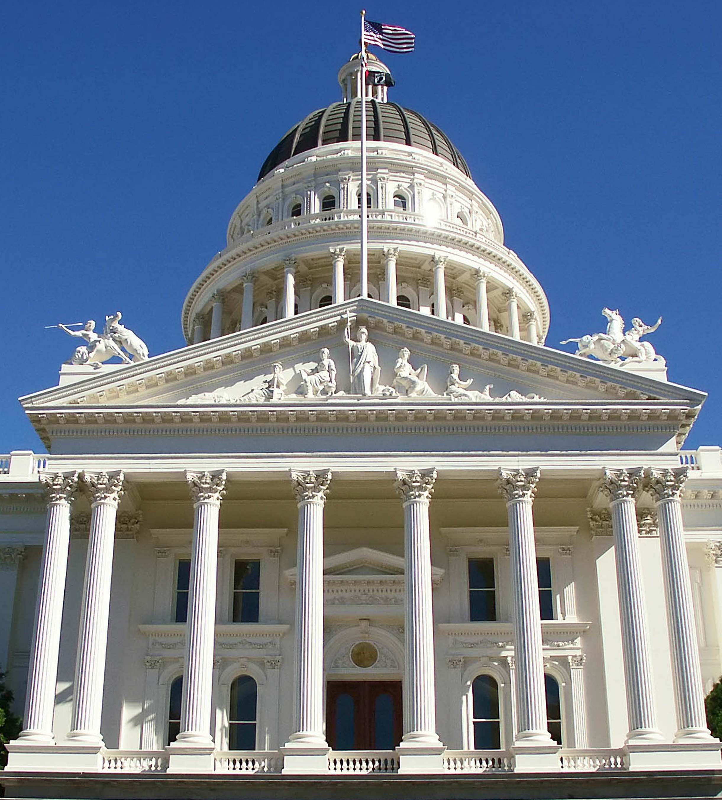 The California State Capitol building, with its classical architecture, tall columns, sculptures on the roof, and an American flag flying on top, against a clear blue sky.