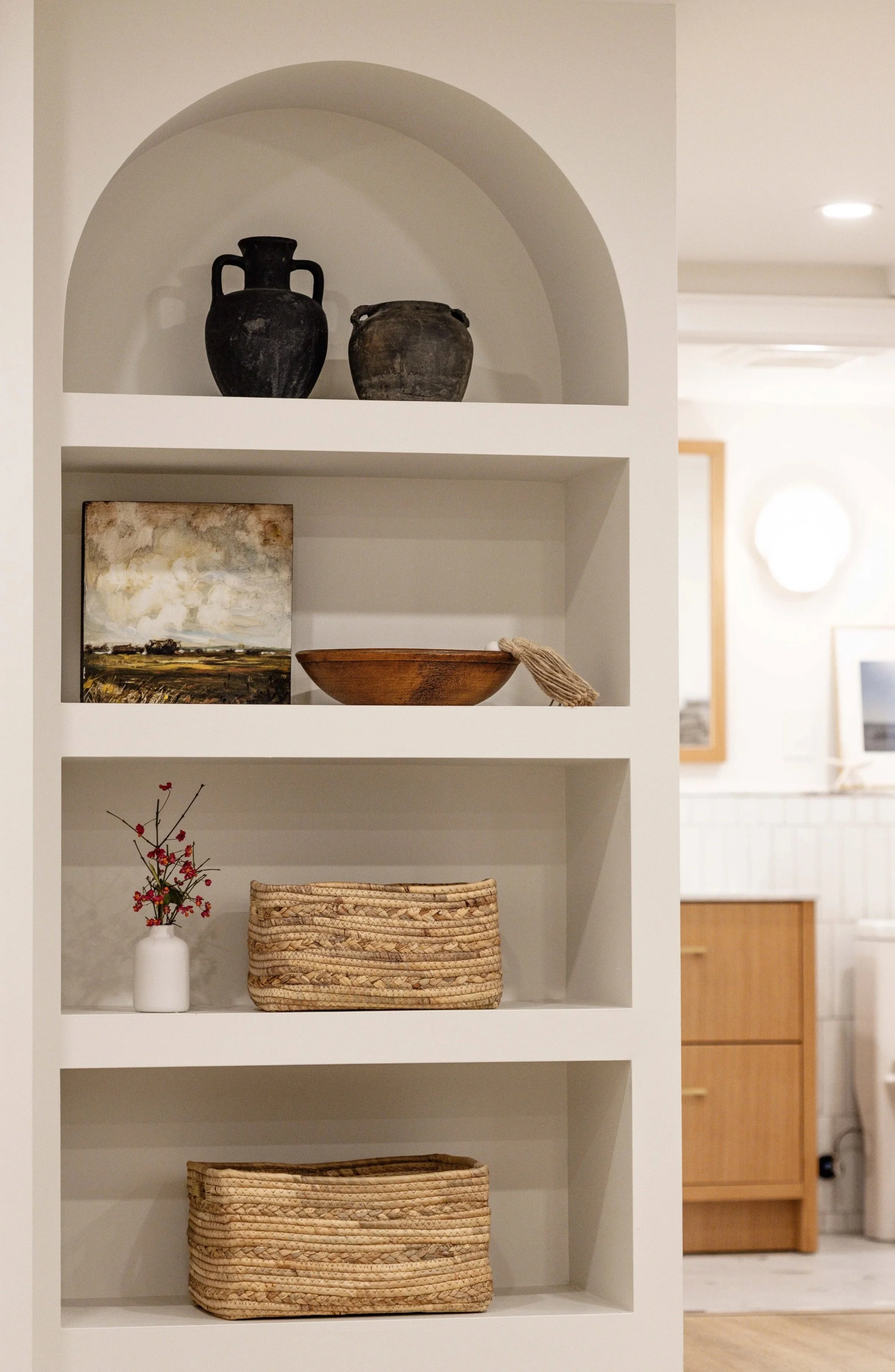 A white built-in shelf with decorative items, including black vases, a landscape painting, a wooden bowl, a small white vase with red berries, and woven baskets, in a room with white walls and wooden furniture.