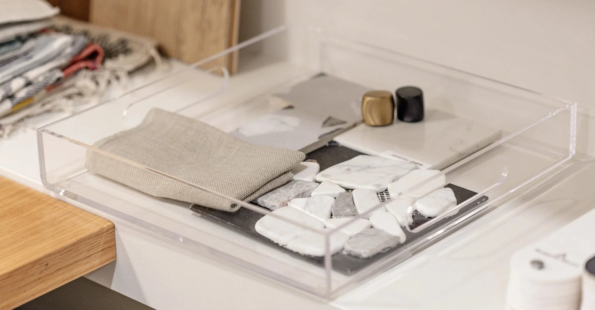 A clear acrylic tray with various stones, fabric swatch, and small decorative objects on a white surface.