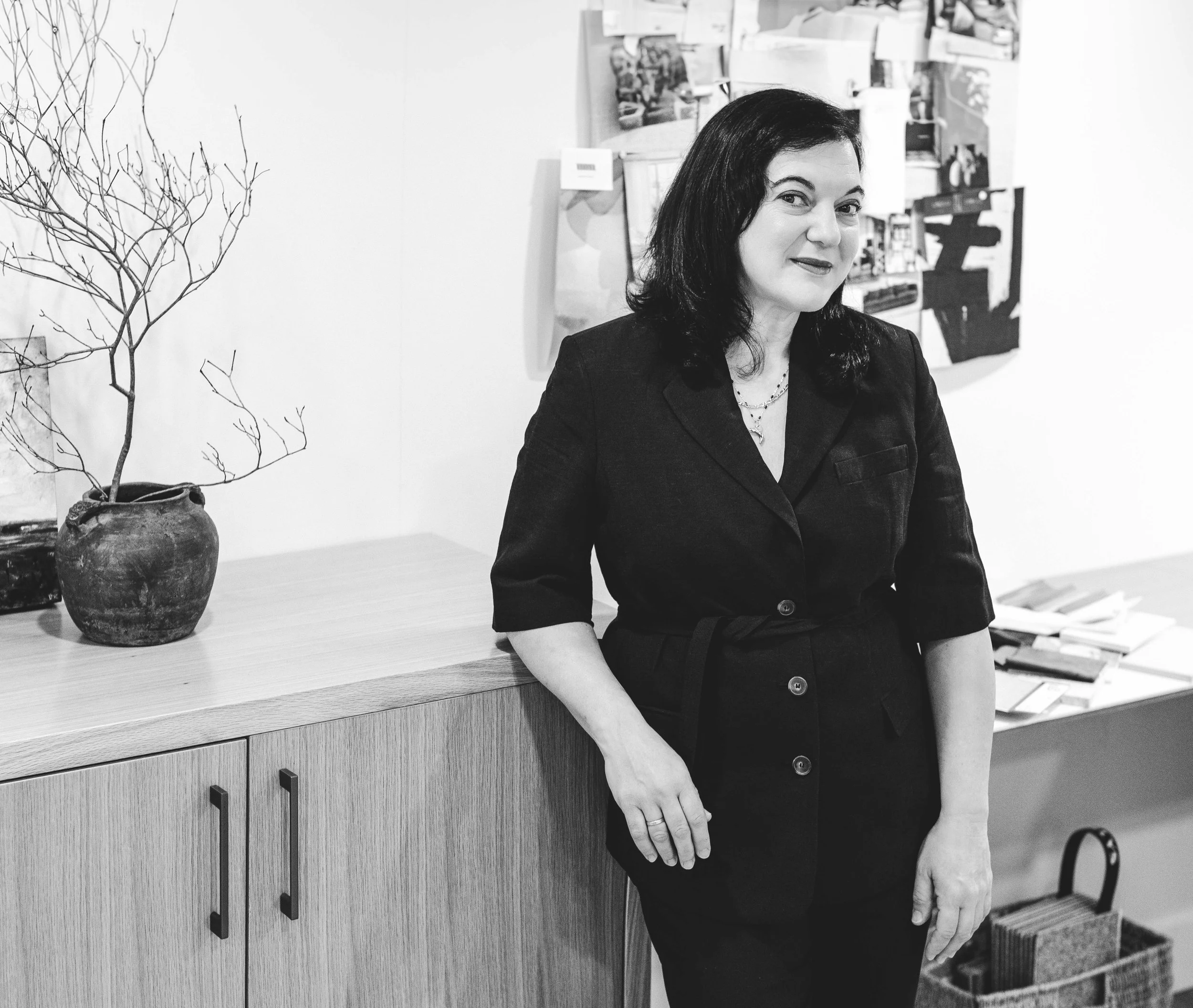 A woman with dark hair wearing a dark blazer and pants, standing next to a wooden cabinet in an art gallery or office, with a paper leafless plant in a clay pot on the cabinet, and wall art and papers in the background.