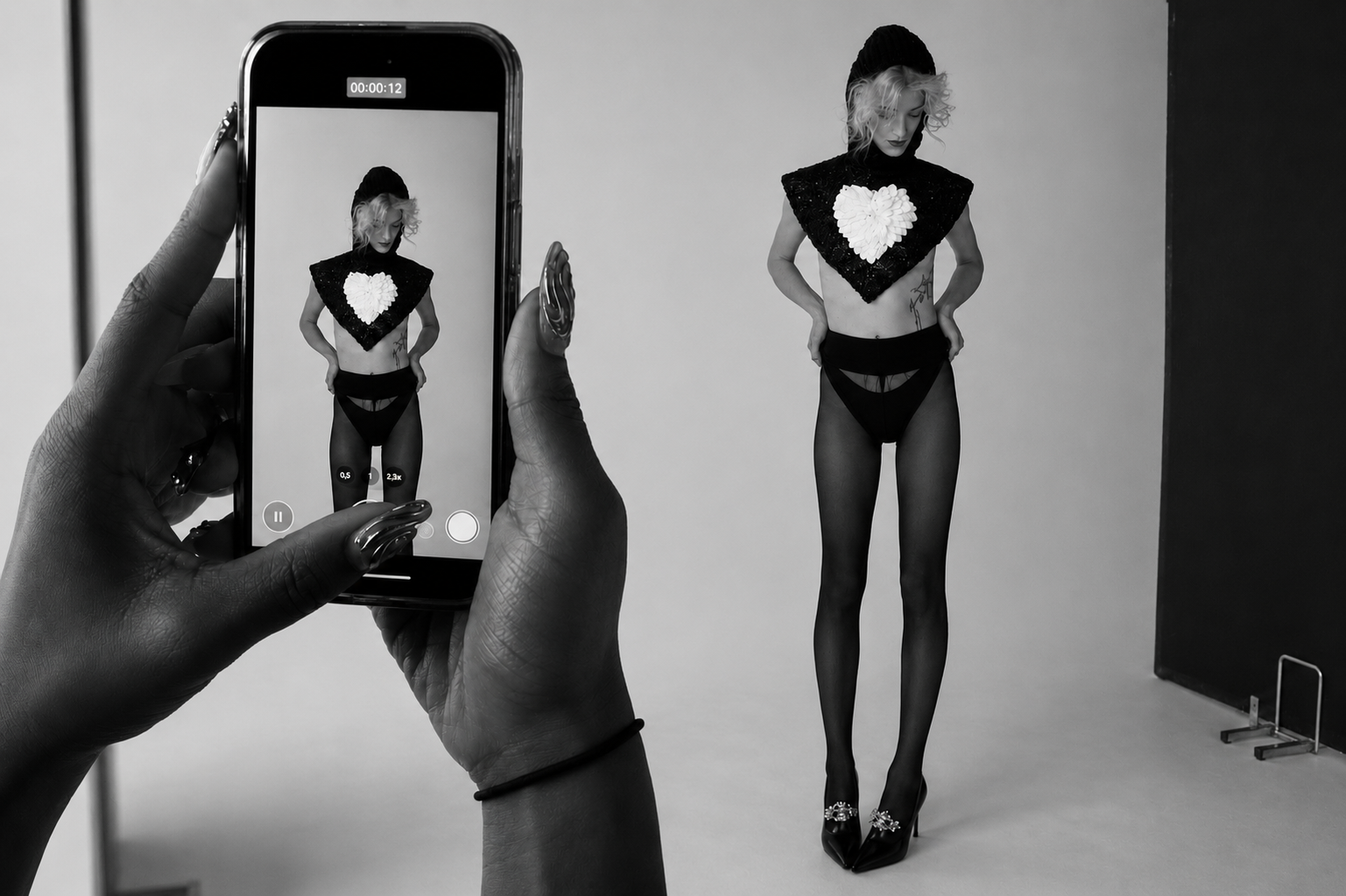 A person taking a black-and-white photo of a model standing against a plain background. The model wears a beanie, a large black triangular top with a white heart design, black stockings, and high heels.