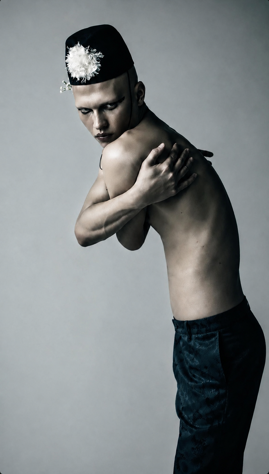 A shirtless man with a pale complexion wearing a black hat with a white feather decoration, striking a dramatic pose with one arm crossing his chest and the other hand resting on his shoulder, against a plain gray background.
