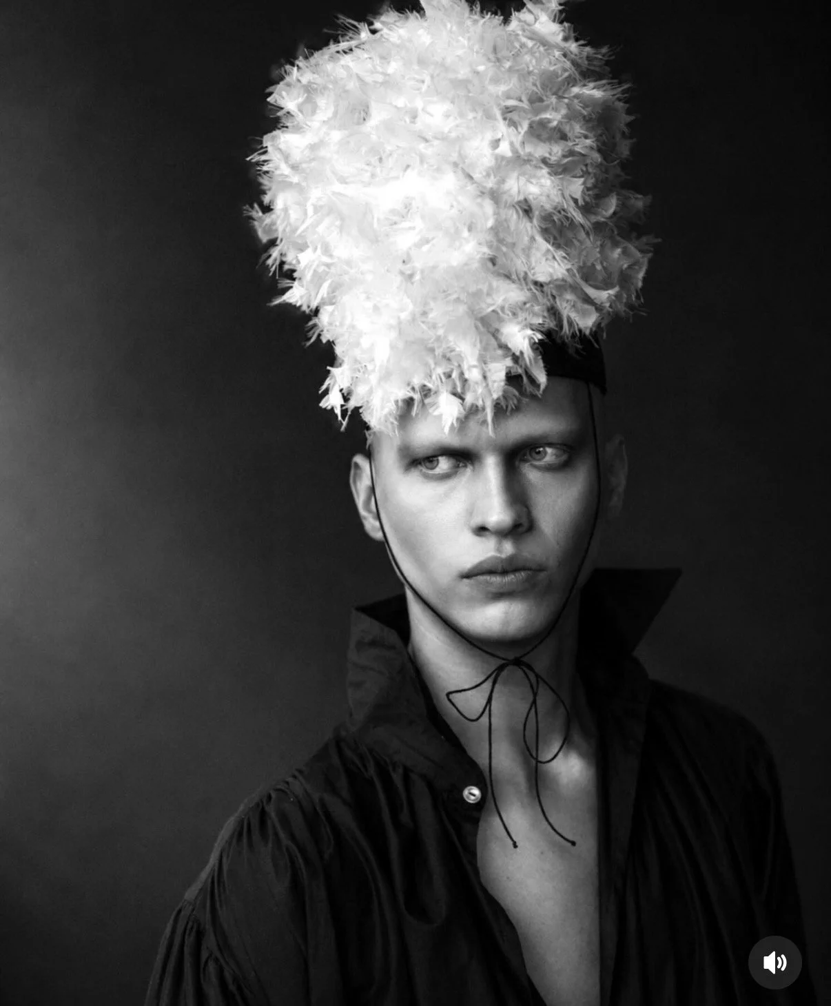 Black and white photo of a person with a serious expression, wearing a feathered headpiece and a dark jacket with an open collar, against a plain backdrop.