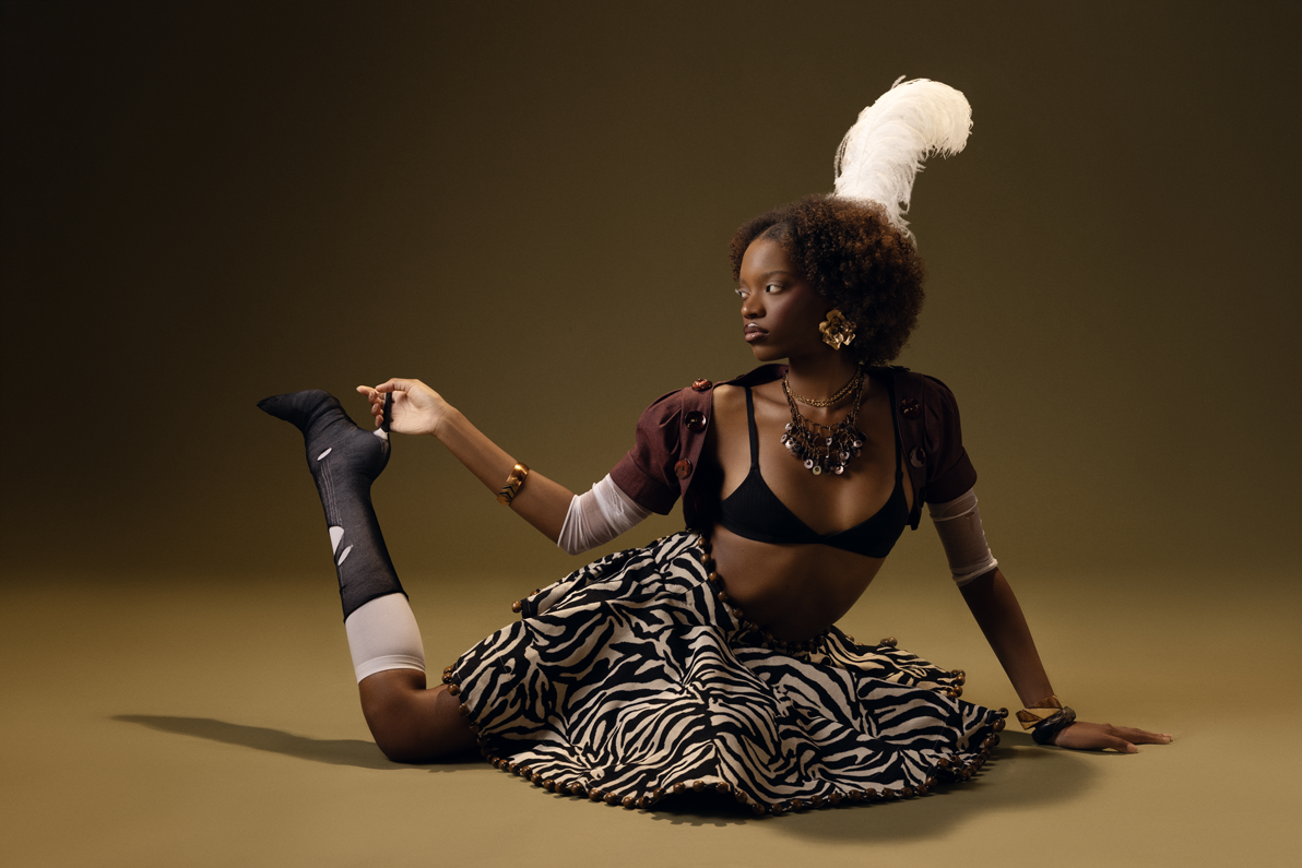 A woman with dark skin and curly hair poses on the ground, wearing a zebra-print skirt, black bra, layered necklaces, earrings, and a feathered headpiece, against a plain dark background.