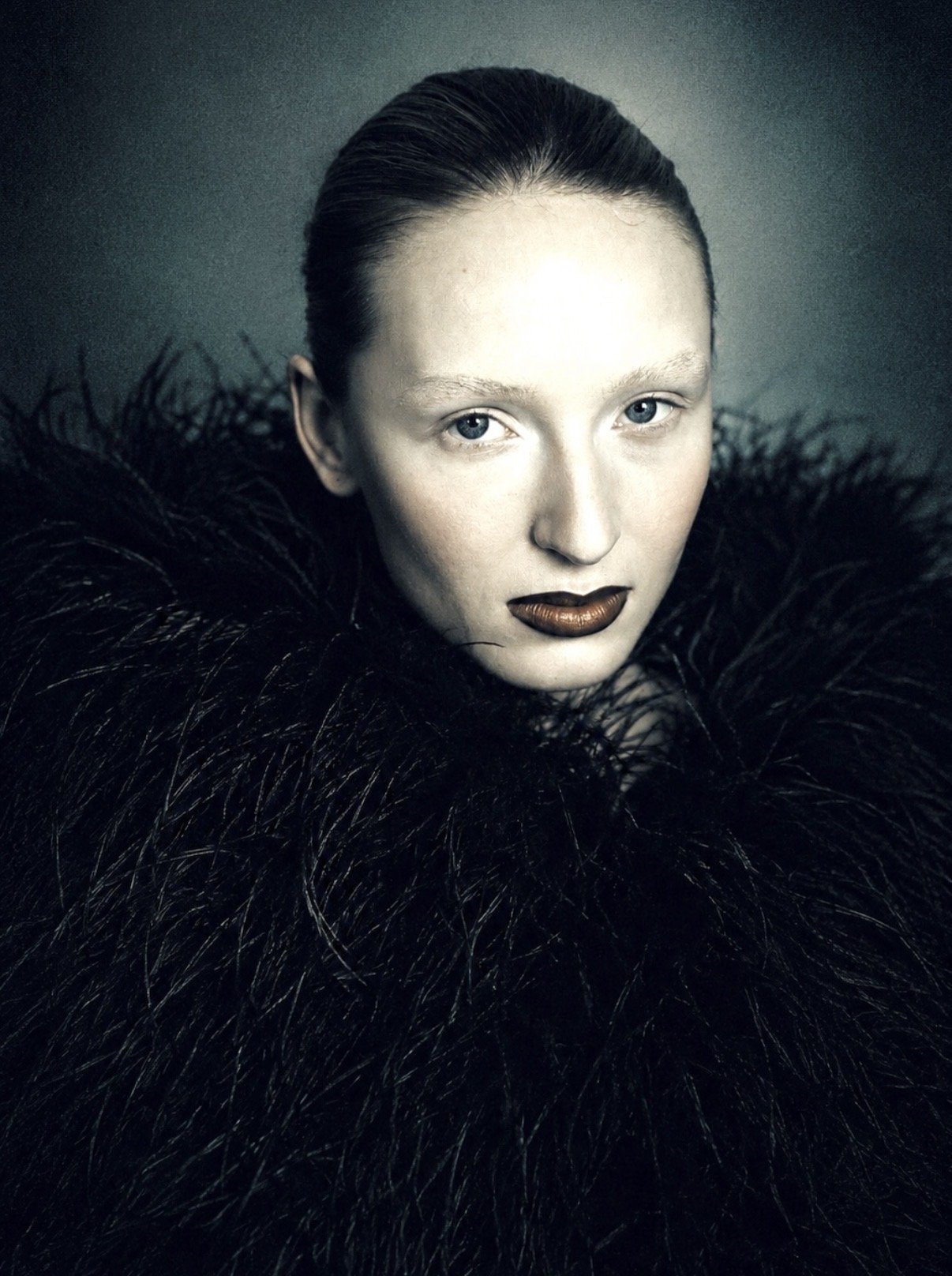 A woman with platinum blonde hair styled back, wearing dark lipstick, with a serious expression, wrapped in a large black feathered garment.