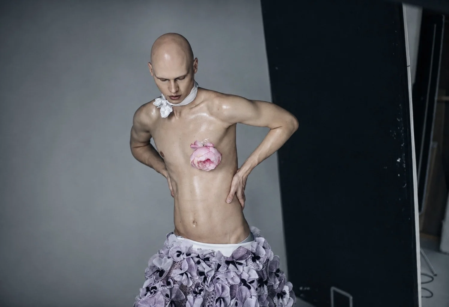 A bald person with a bare upper body and a pink flower covering their chest, wearing a white scarf around their neck, and a skirt made of multiple layered ruffled fabric. They are standing against a gray background, looking down with one hand on thei
