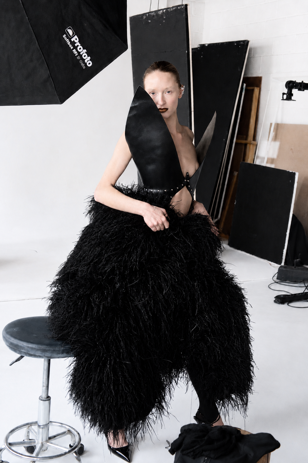 A woman with makeup and slicked-back hair wearing a black feathered skirt and large black leather wings posed in a photography studio.