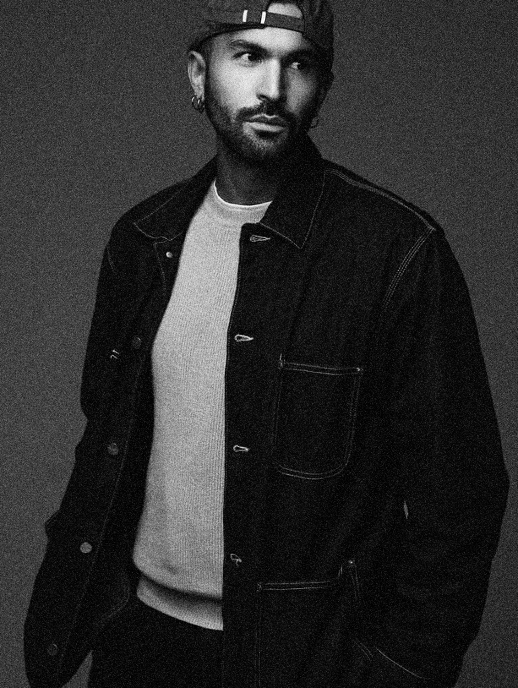 Black and white portrait of a man with dark hair, beard, and earrings, wearing a backward baseball cap, denim jacket, and a light-colored shirt, looking to the side, against a plain background.