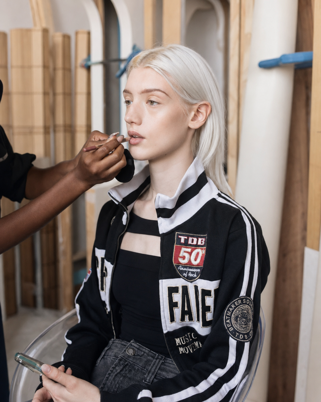 A young woman with platinum blonde hair sitting in a makeup chair while a makeup artist applies makeup to her lips, inside a studio with wooden walls and equipment.