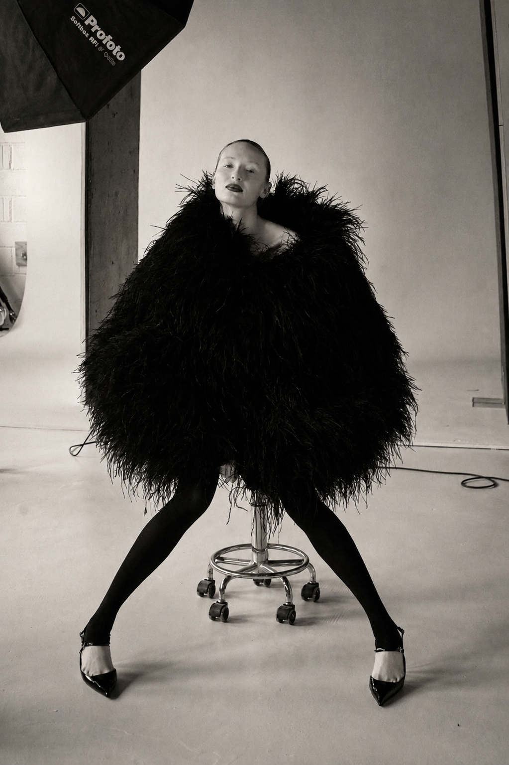 A woman sitting on a rolling stool in a photography studio, wearing a large black feathered coat, black tights, and high heels. She has a confident pose with legs apart and head turned, with visible studio equipment in the background.