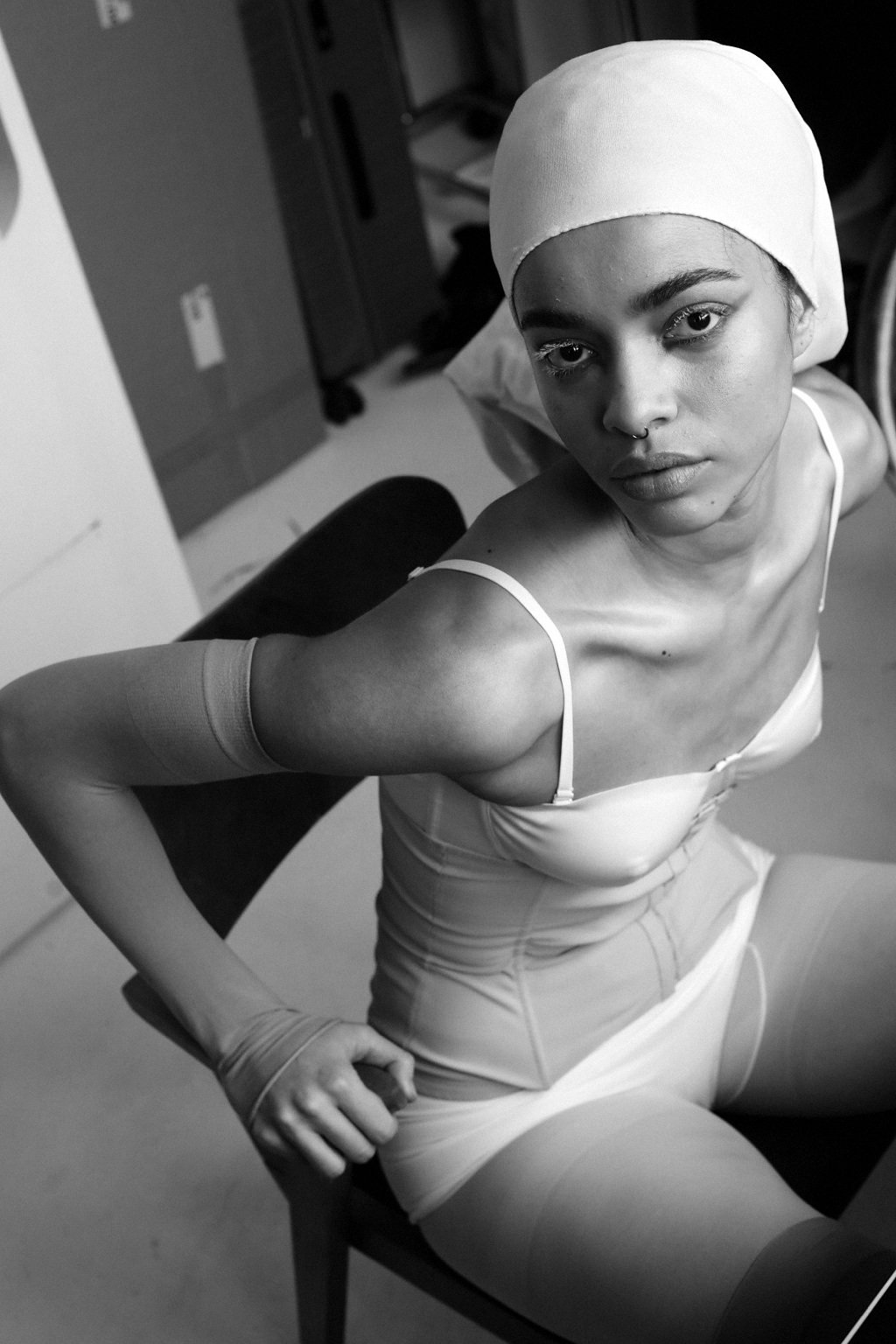 A young woman with short hair wearing a white headscarf, a spaghetti strap top, and shorts, sitting on a chair in an indoor setting.