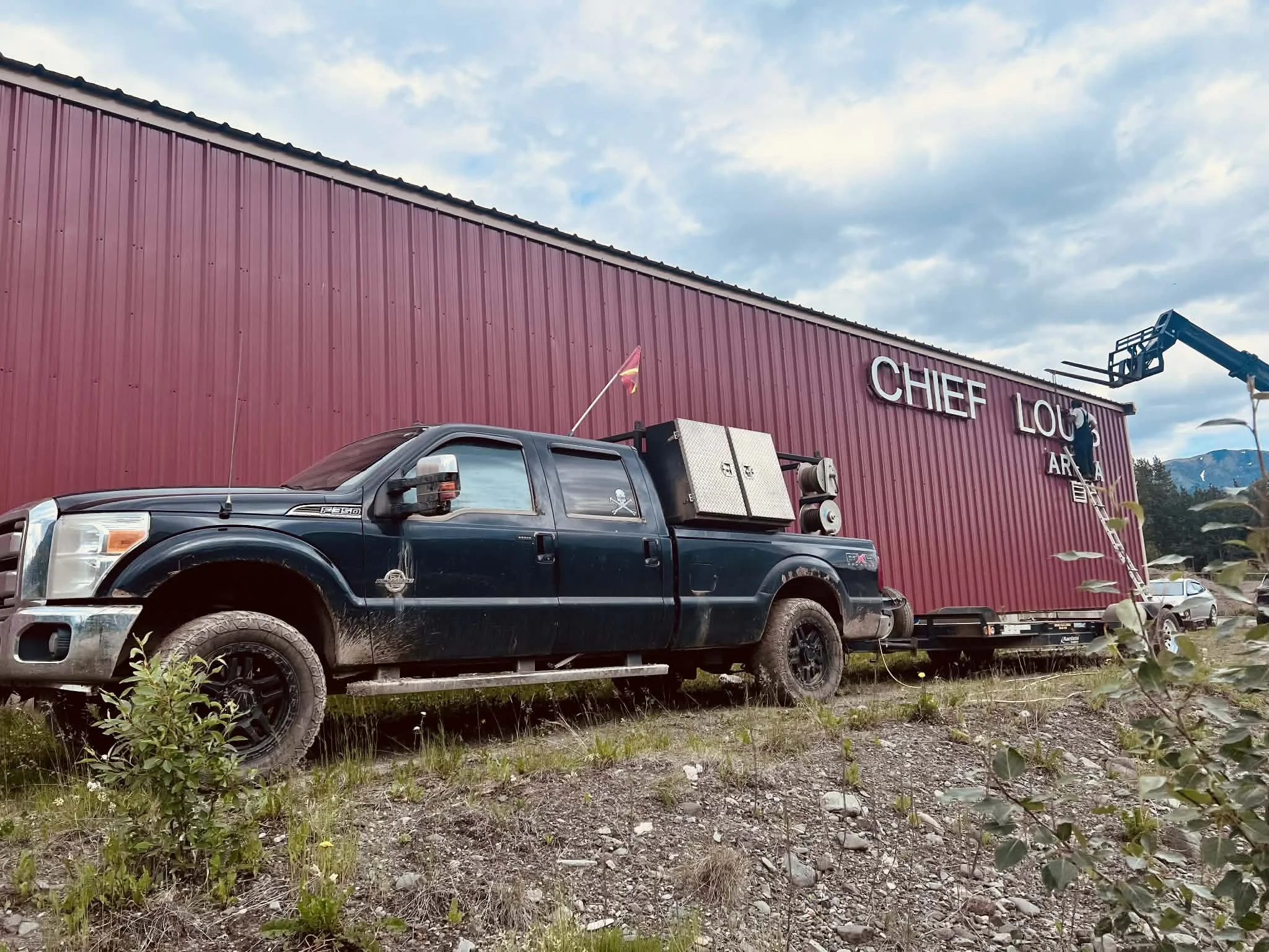 A black pickup truck parked on a patch of dirt and grass in front of a large red building with the words 'Chief Lou' on it. The truck has equipment in the back and a small flag attached to the side mirror. There is a cherry picker lift extended to the building and a person working on the sign.