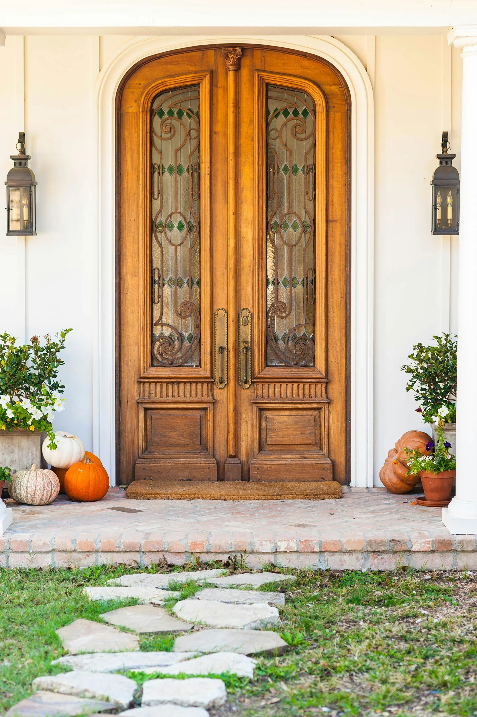 Wooden front door with decorative glass panels, surrounded by outdoor lanterns, pumpkins, and potted plants, with stone pathway leading up to it.