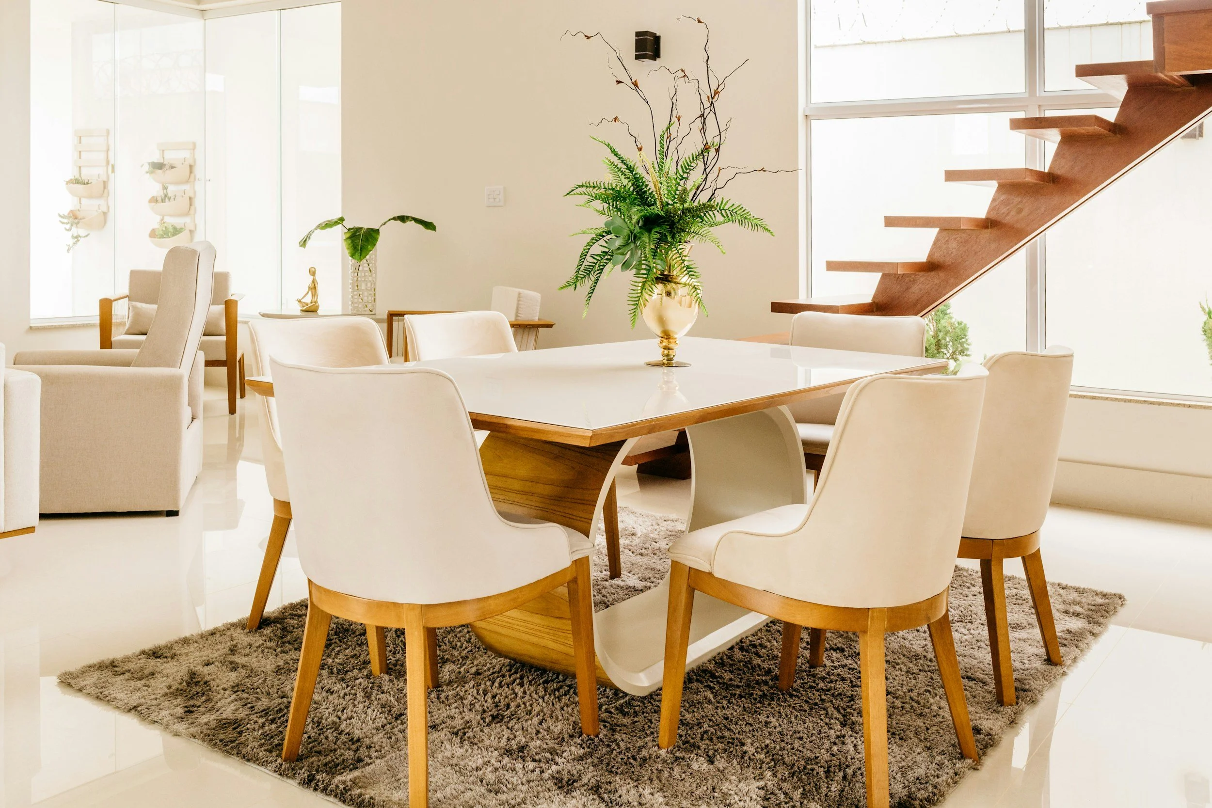Modern dining room with a white table, cream chairs, a large green potted fern, wooden staircase, and large windows.