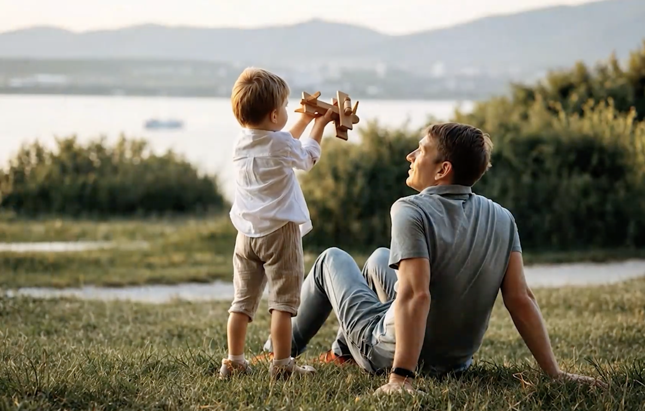 Father and young son playing with a wooden toy plane on the grass by the lake at sunset at Liberty Hills