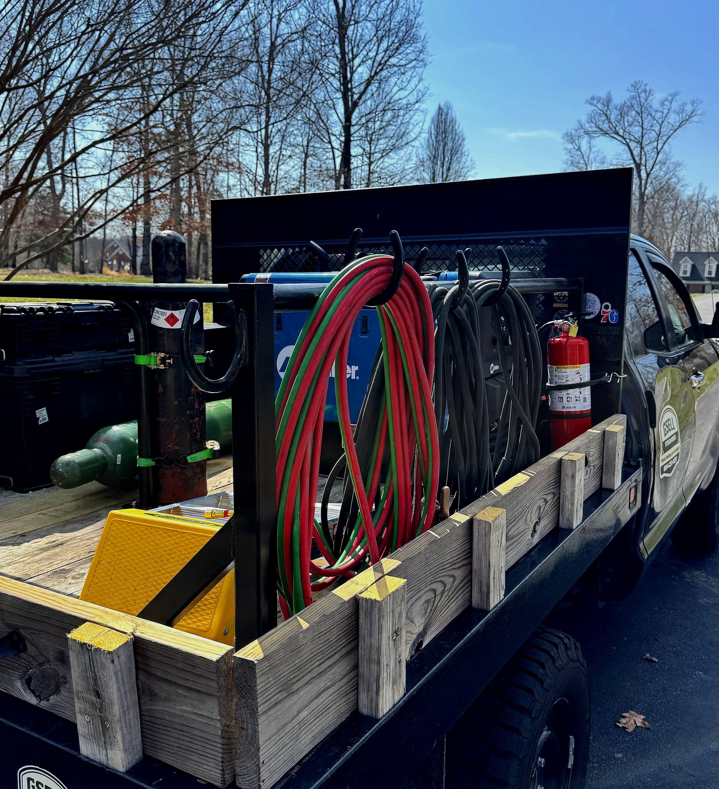 Utility truck bed with wood sides, carrying assorted equipment including welding cables, a fire extinguisher, and various welding tools, set outdoors with trees and houses in the background on a clear day.