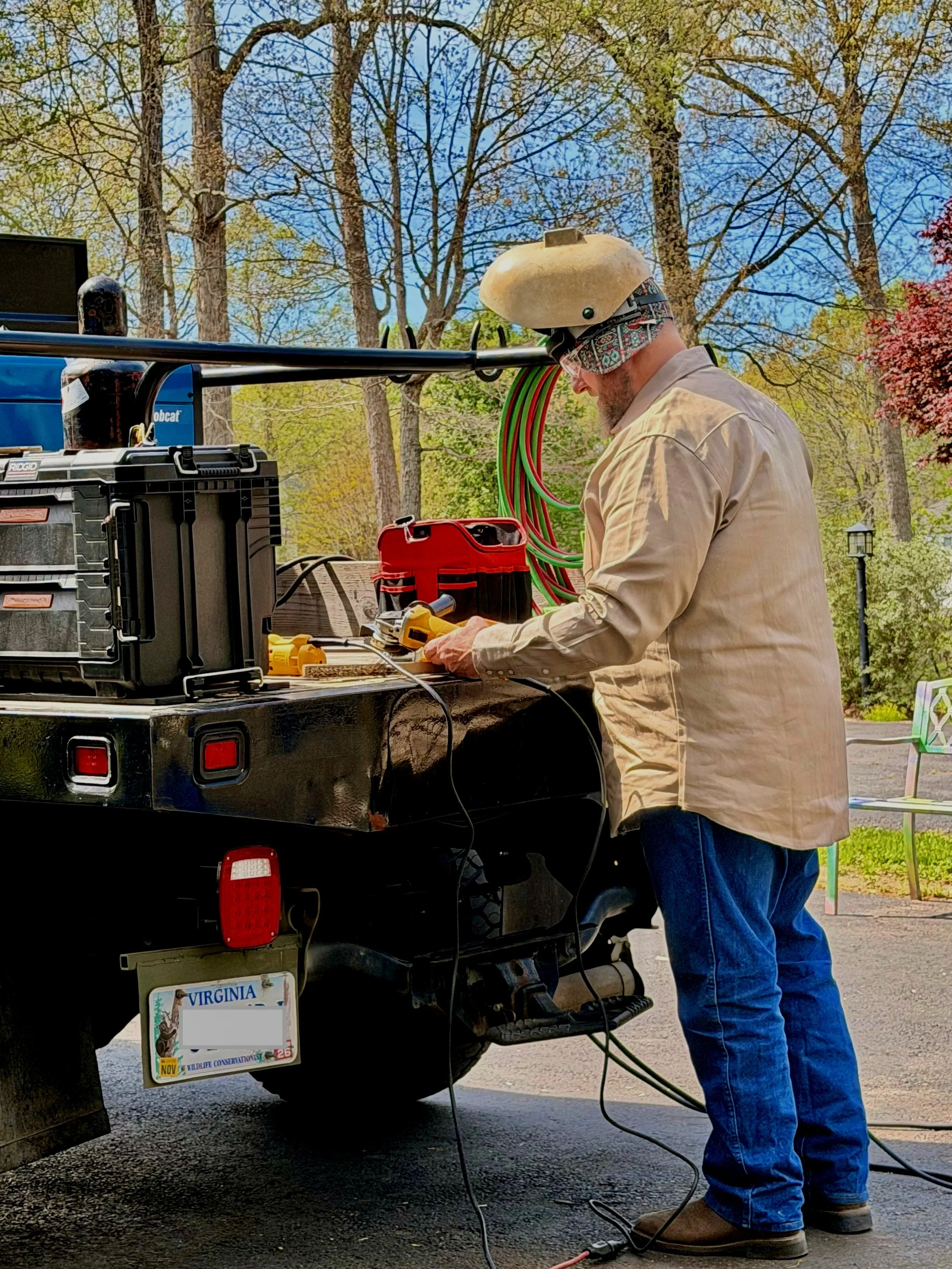 A man in a beige shirt, blue jeans, and brown boots wearing a helmet and eye protection working with electrical equipment on the back of a black pickup truck. The truck has a Virginia license plate and various electrical wires and tools are on the truck bed. There are trees with green leaves and a lamppost in the background.