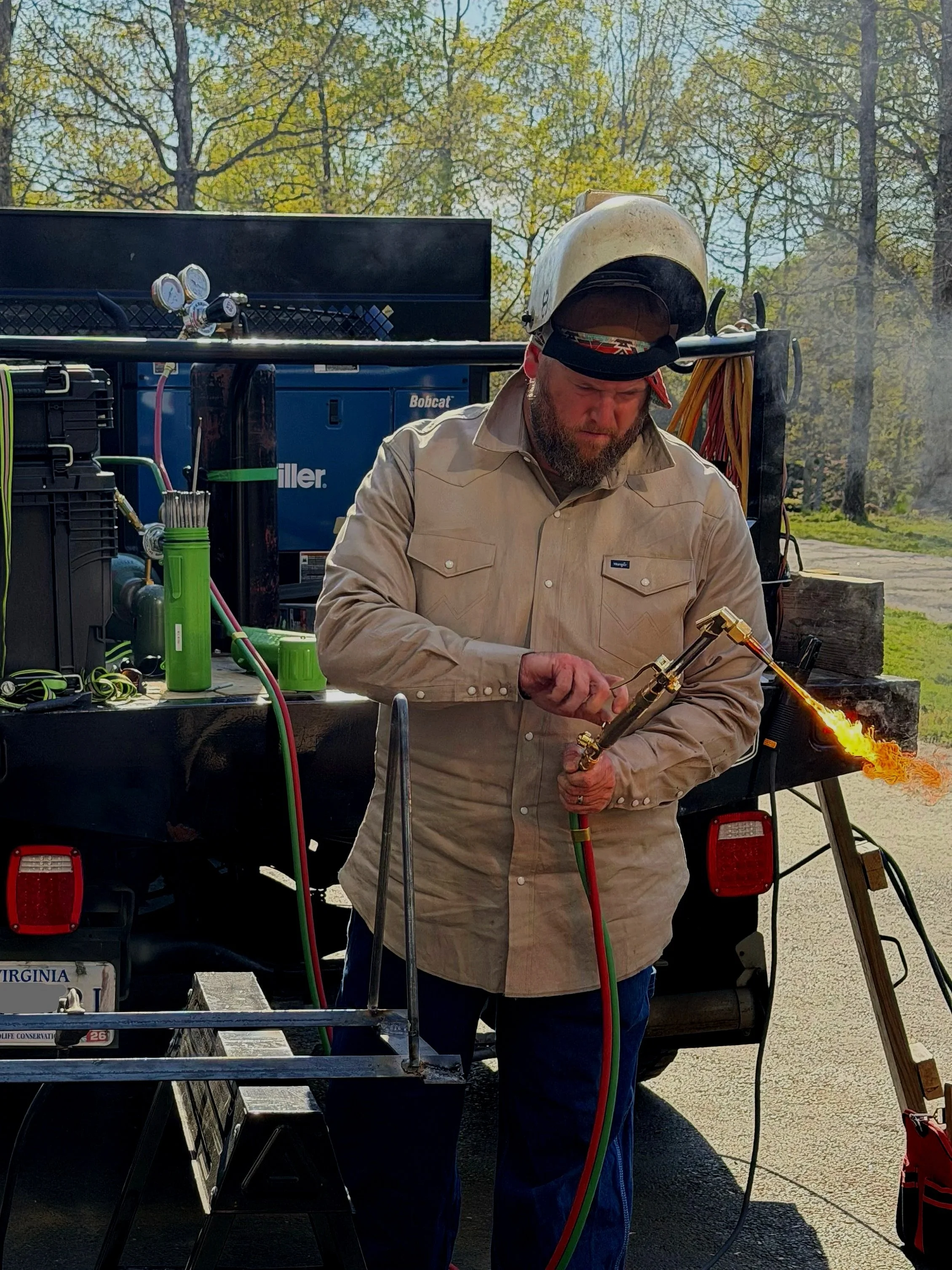 A man wearing a welding helmet and beige shirt using a cutting torch outdoors during daytime, with trees and a blue sky in the background.