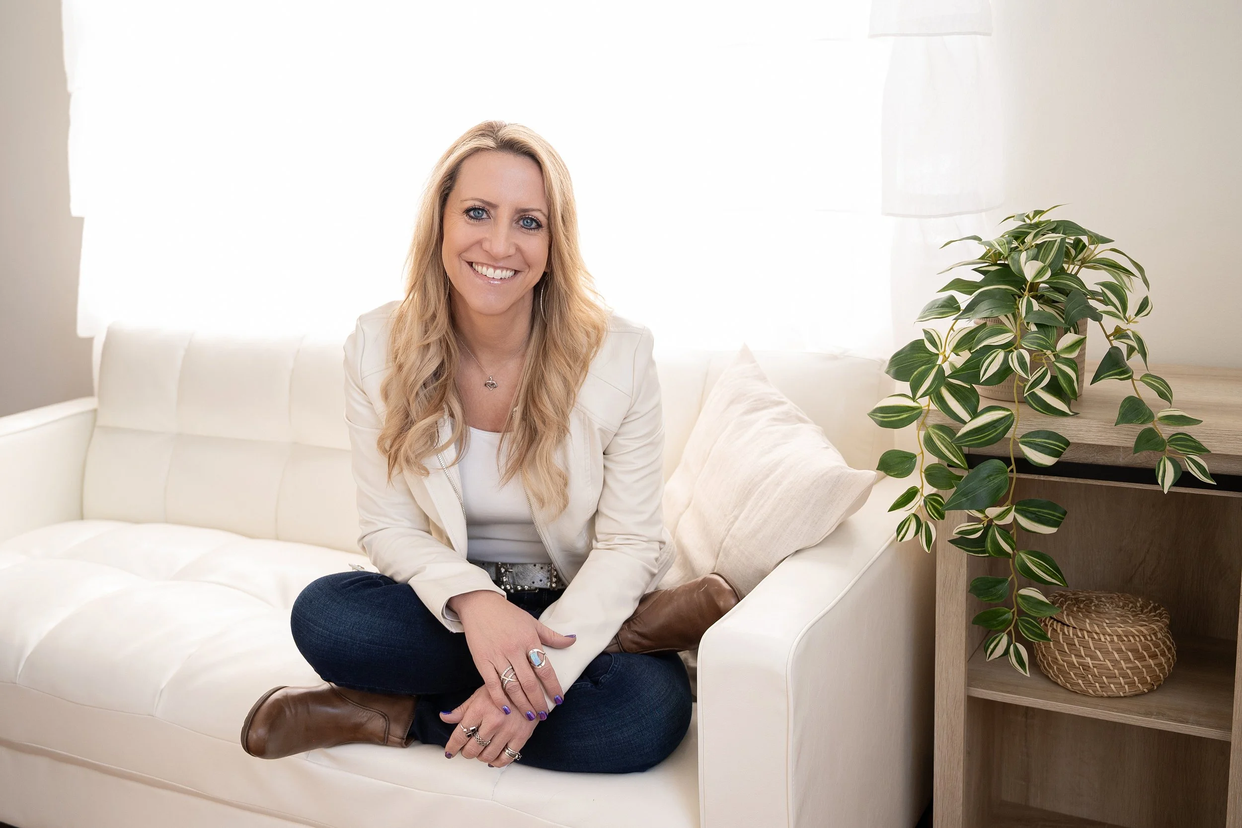 Amy in a white blazer and jeans, sitting on a white couch with the sunlight behind her, smiling