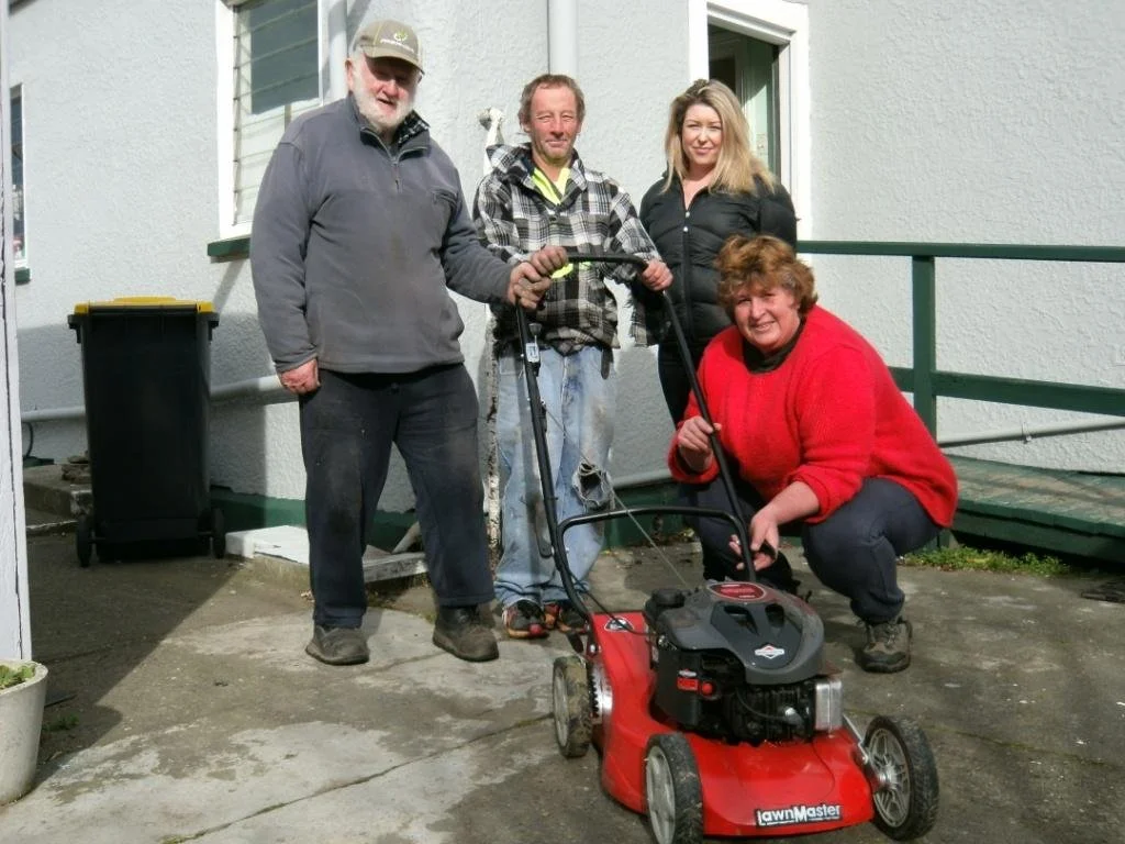 Four people outside near a white building with green accents, with a lawnmower. Two men and two women, one woman in red crouching, and the others standing.
