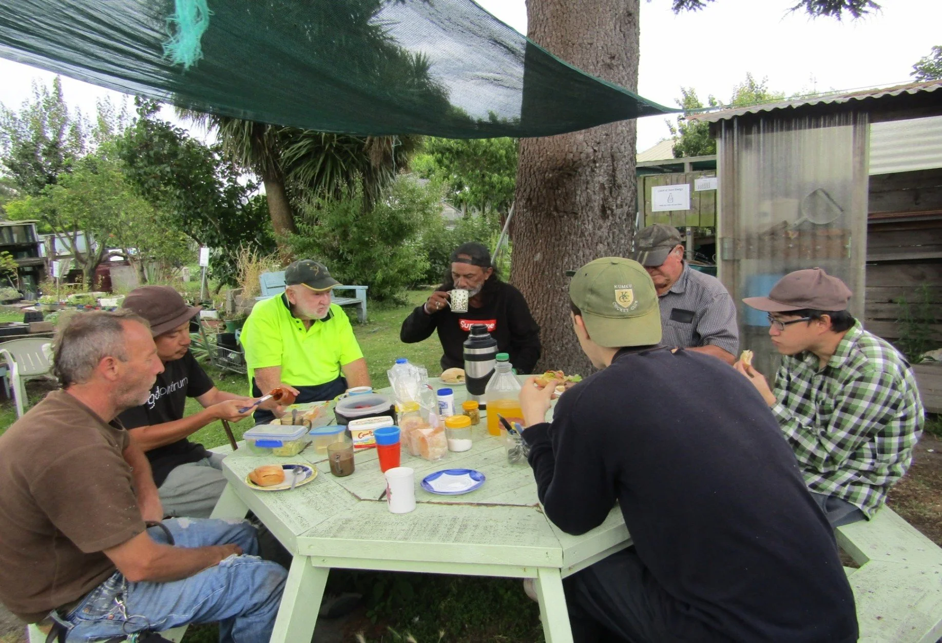 A group of eight people sitting around a outdoor table sharing a meal in a garden, with trees and a shed in the background.