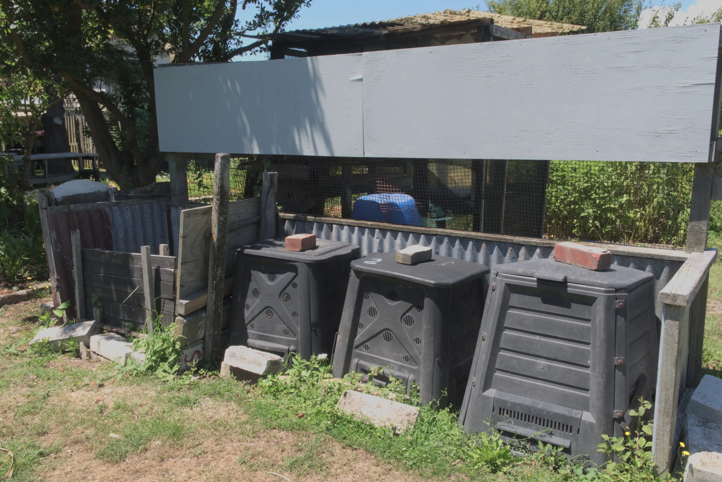 A backyard compost bin area with three black compost bins topped with bricks, in front of a raised garden bed and a makeshift wooden shelter. The area is surrounded by grass and some weeds.