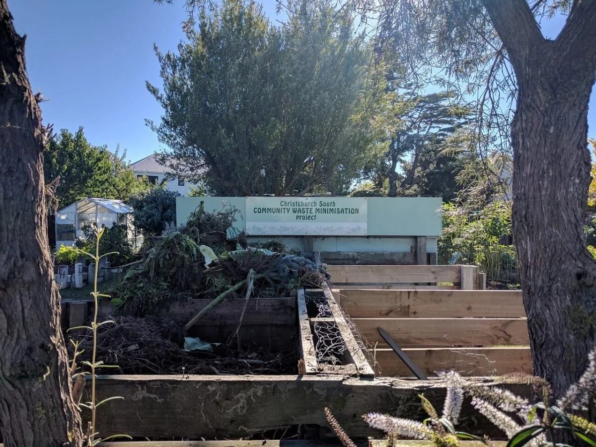 Community garden with compost and waste minimization project sign, surrounded by trees and plants.