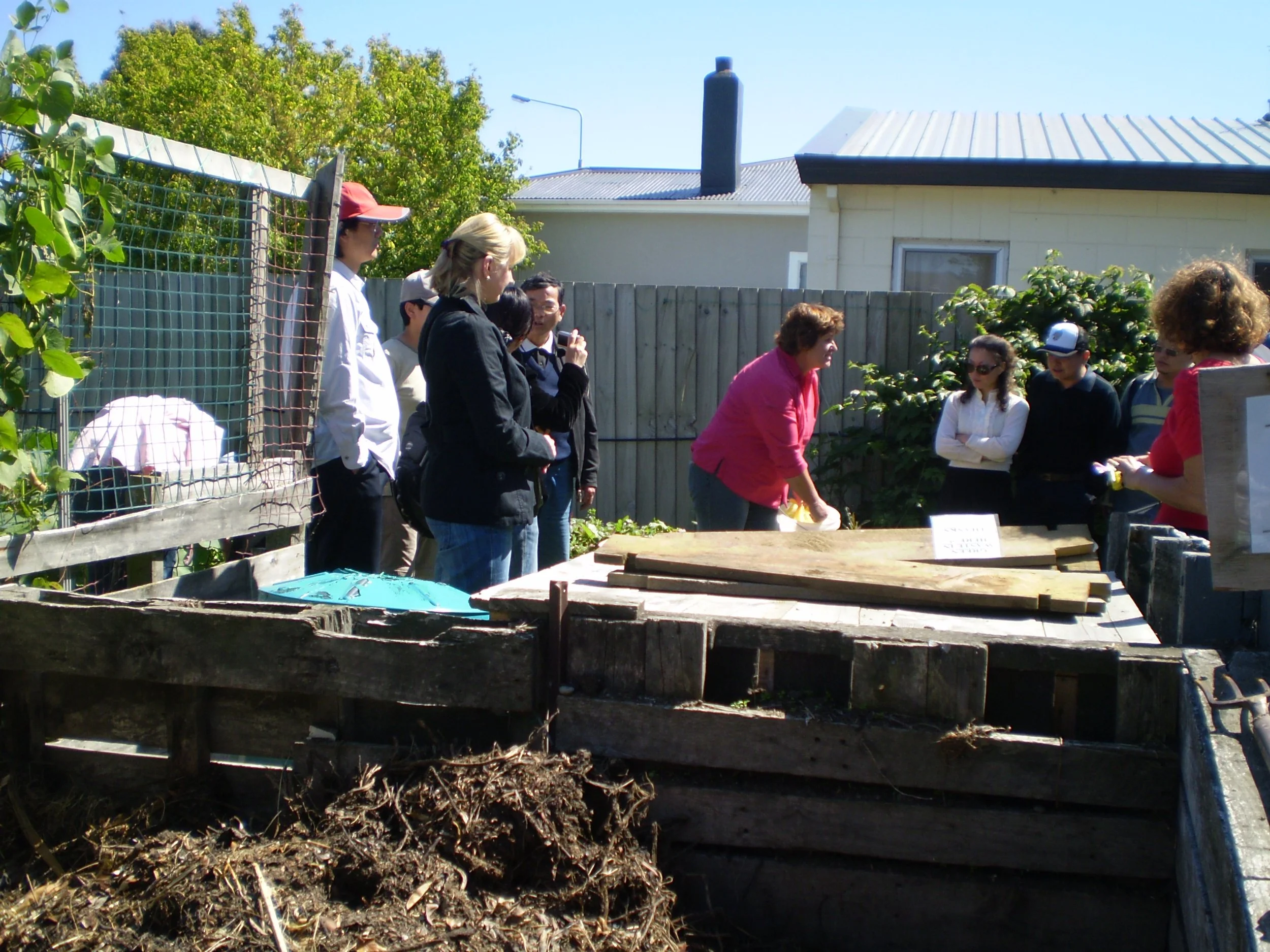Group of people gathered in a backyard garden, some watching as a woman demonstrates planting techniques. The scene is sunny with a wooden fence, trees, and a house in the background.